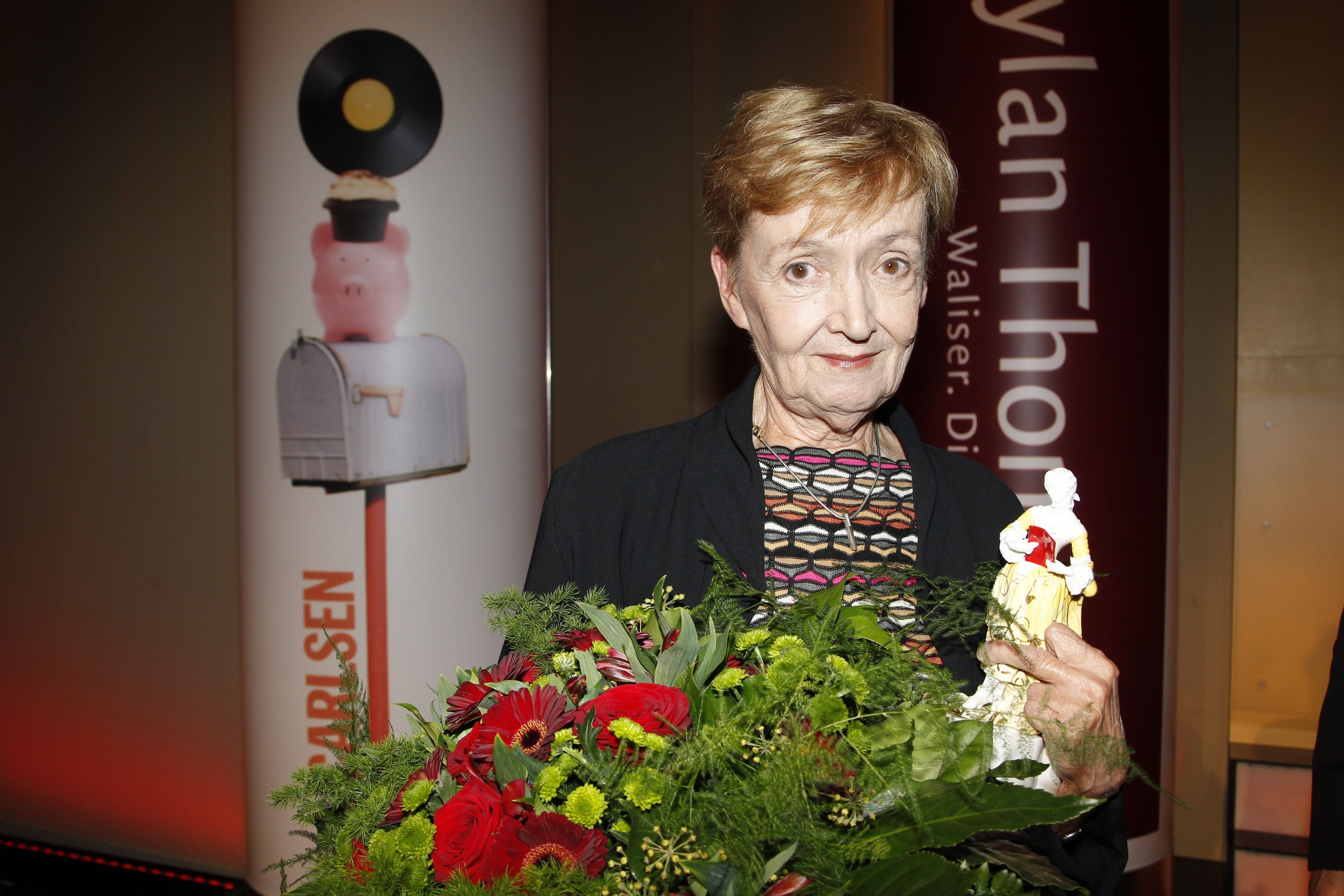 Christine Nöstlinger hält einen Blumenstrauß und eine Figur bei einer Preisverleihung in München.