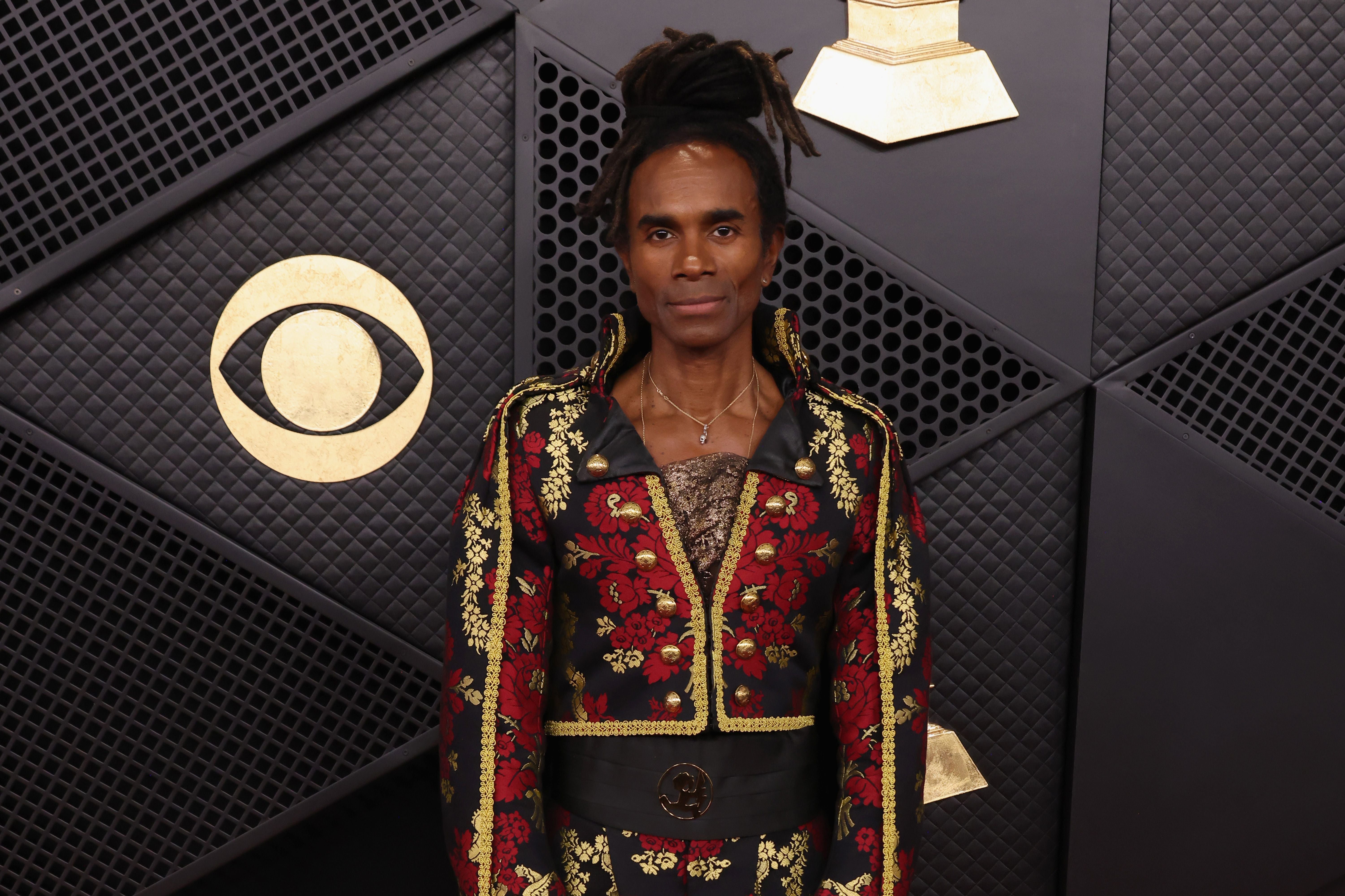 Eine Person mit Dreadlocks trägt einen auffälligen, roten und goldenen Anzug vor GRAMMY-Dekor.