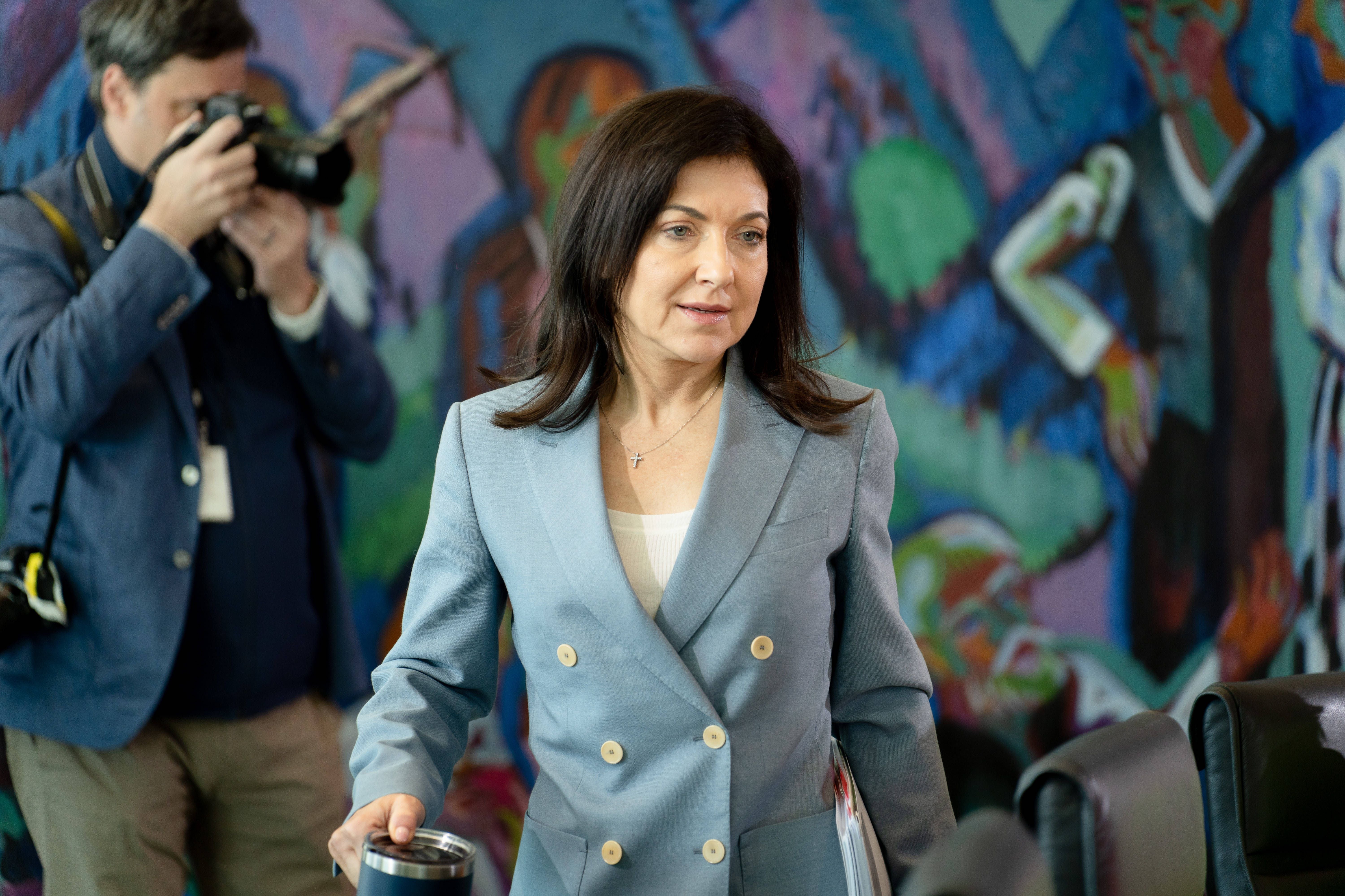 Eine Frau im hellblauen Blazer steht in einem Raum mit buntem Wandbild, im Hintergrund ein Fotograf.