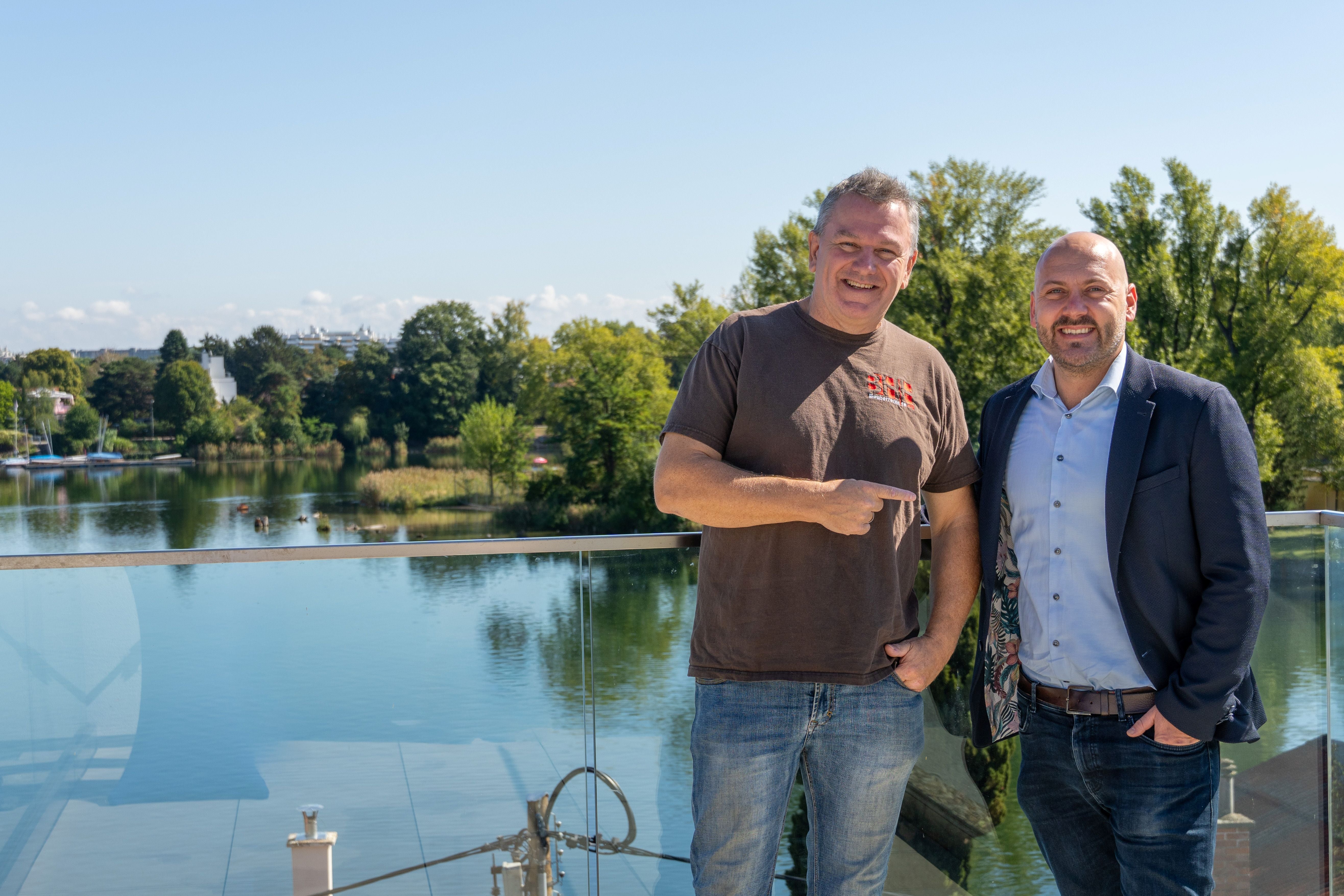 Zwei Männer stehen lächelnd auf einem Balkon mit Blick auf einen ruhigen Fluss und grüne Bäume.