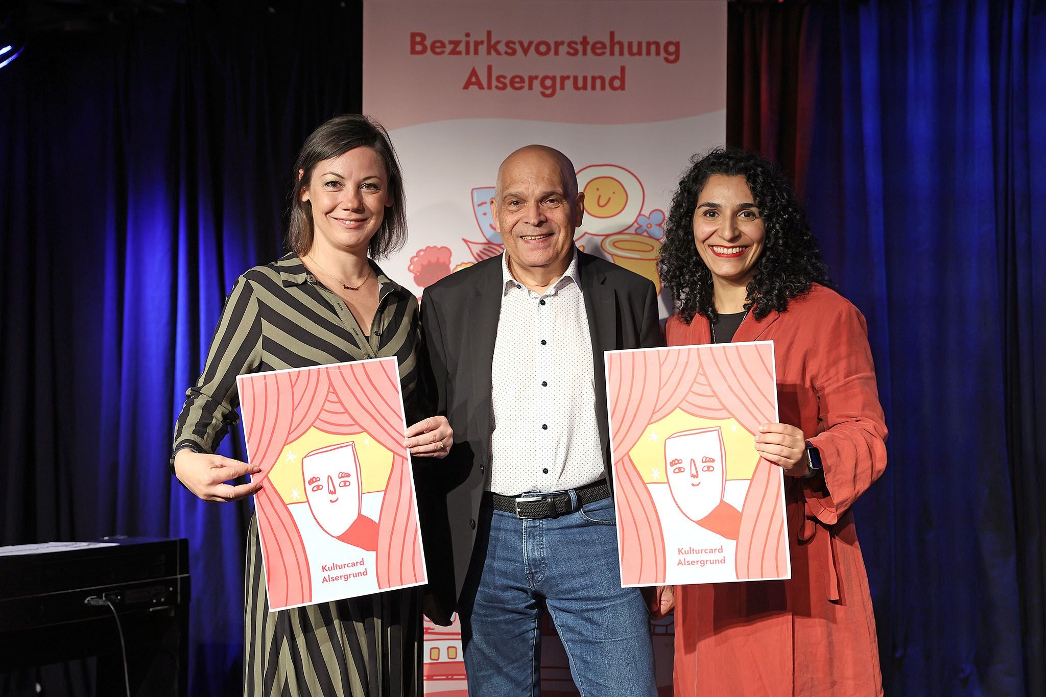 Drei Personen posieren auf einer Bühne mit Plakaten der Kulturcard Alsergrund.