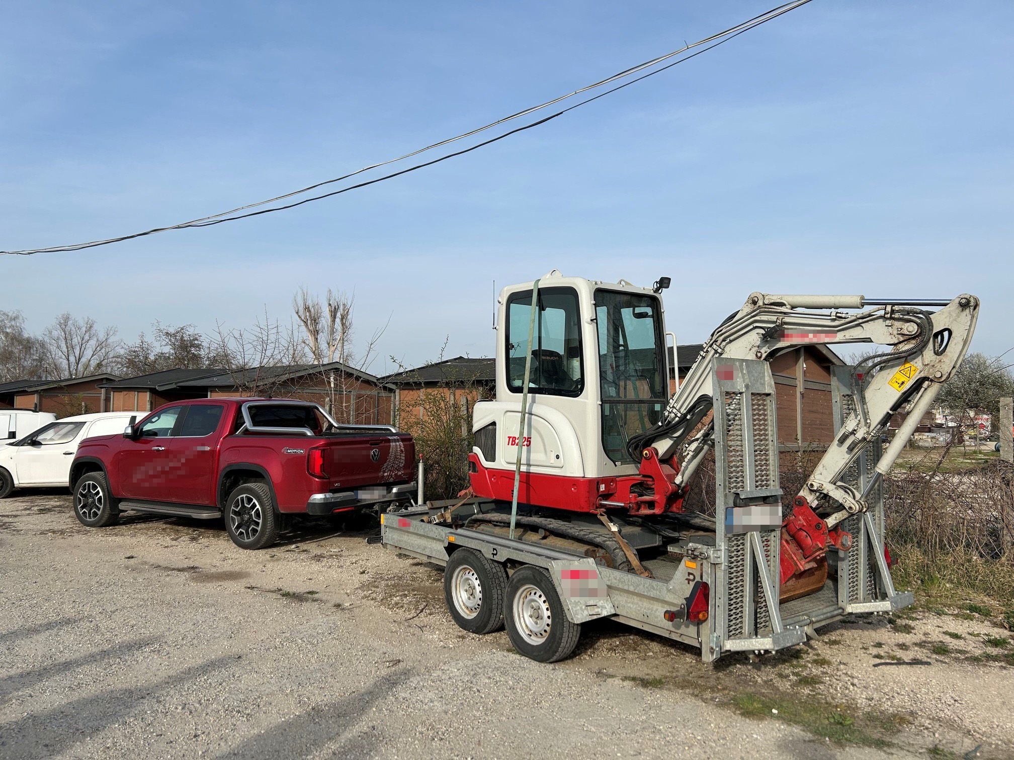Roter Pickup zieht Anhänger mit kleinem Bagger auf einem Parkplatz vor Gebäuden.