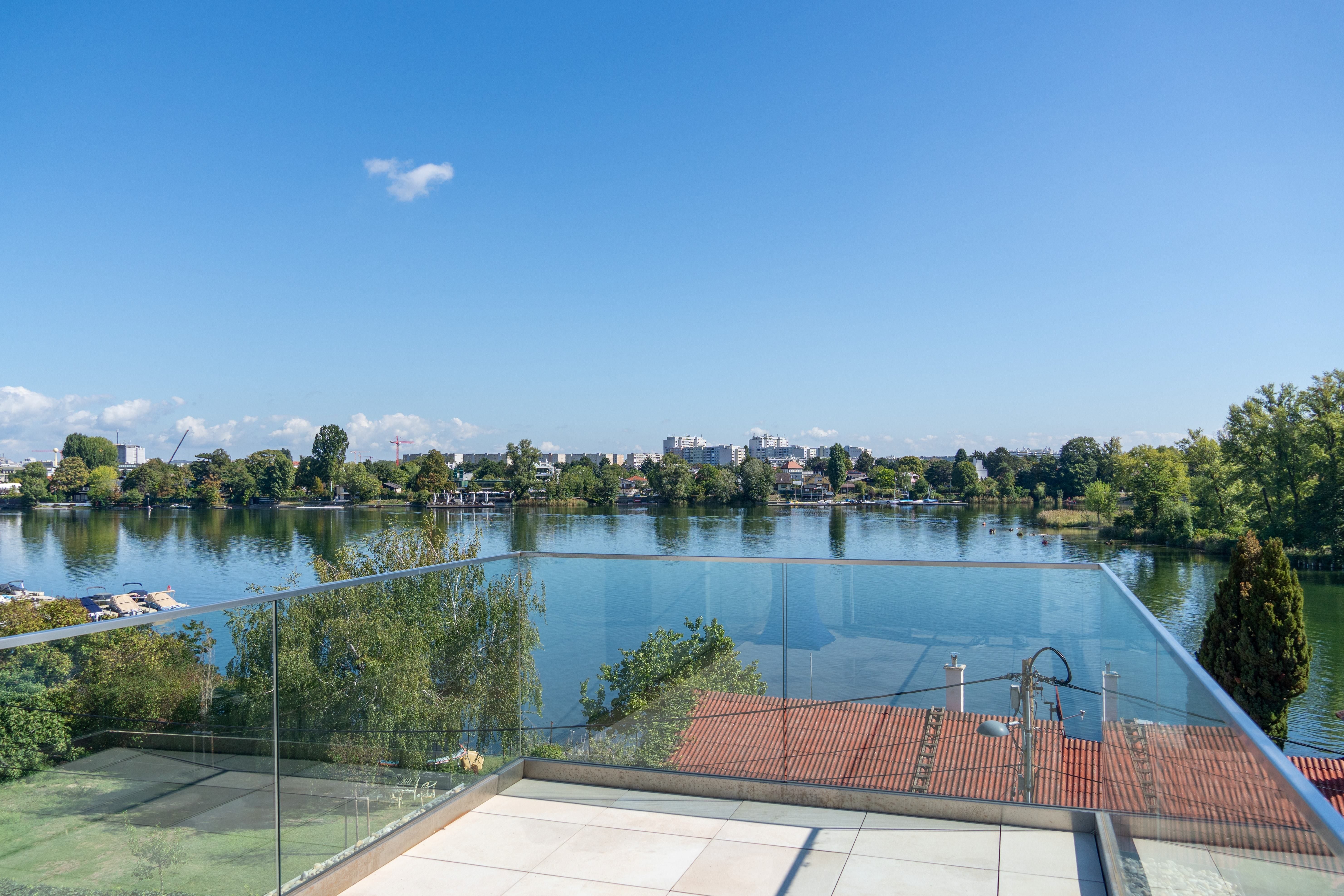 Blick von einer Terrasse mit Glasgeländer auf einen Fluss und grüne Landschaft bei klarem Himmel.