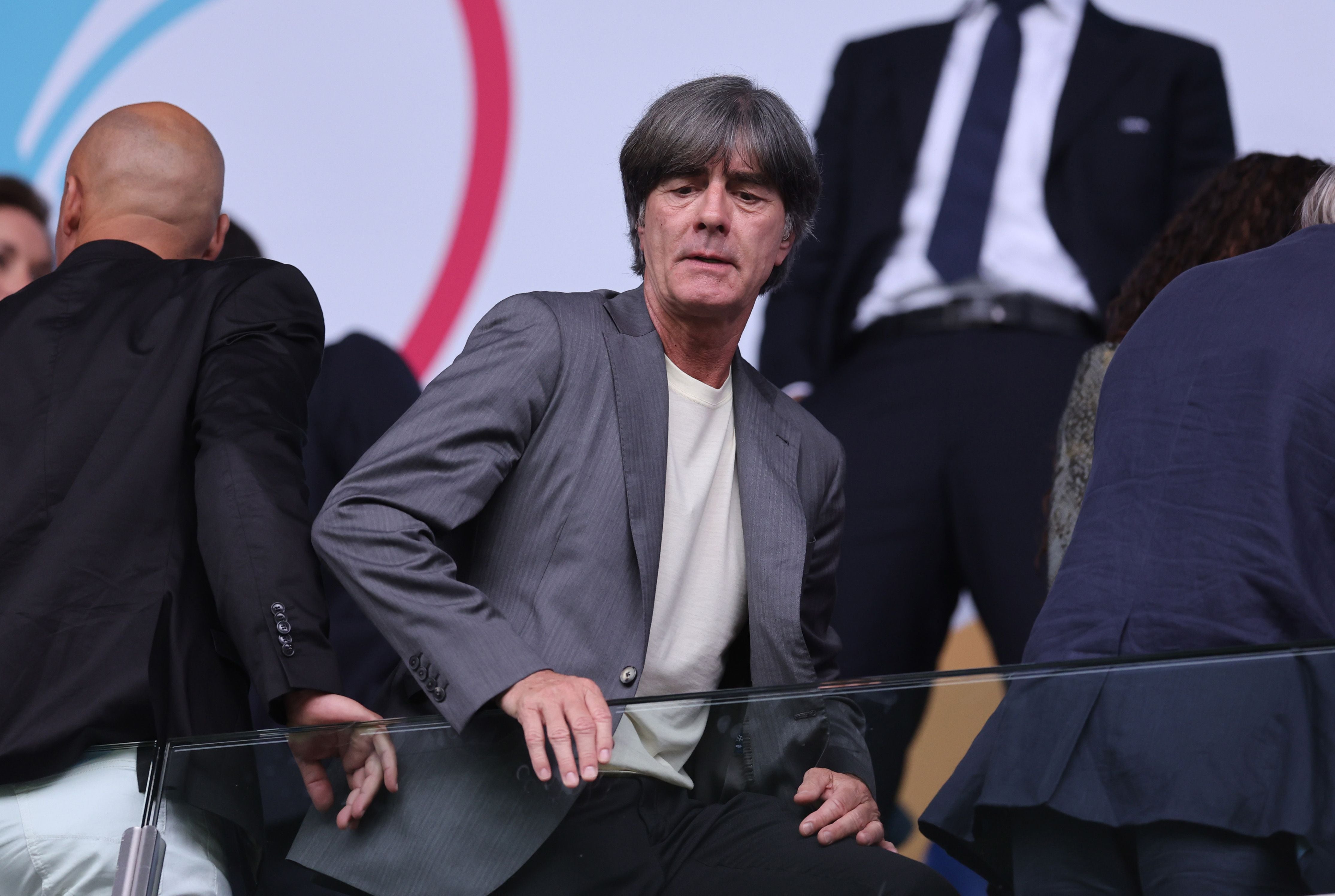 Ein Mann im grauen Anzug sitzt in einem Stadion mit Menschen im Hintergrund.