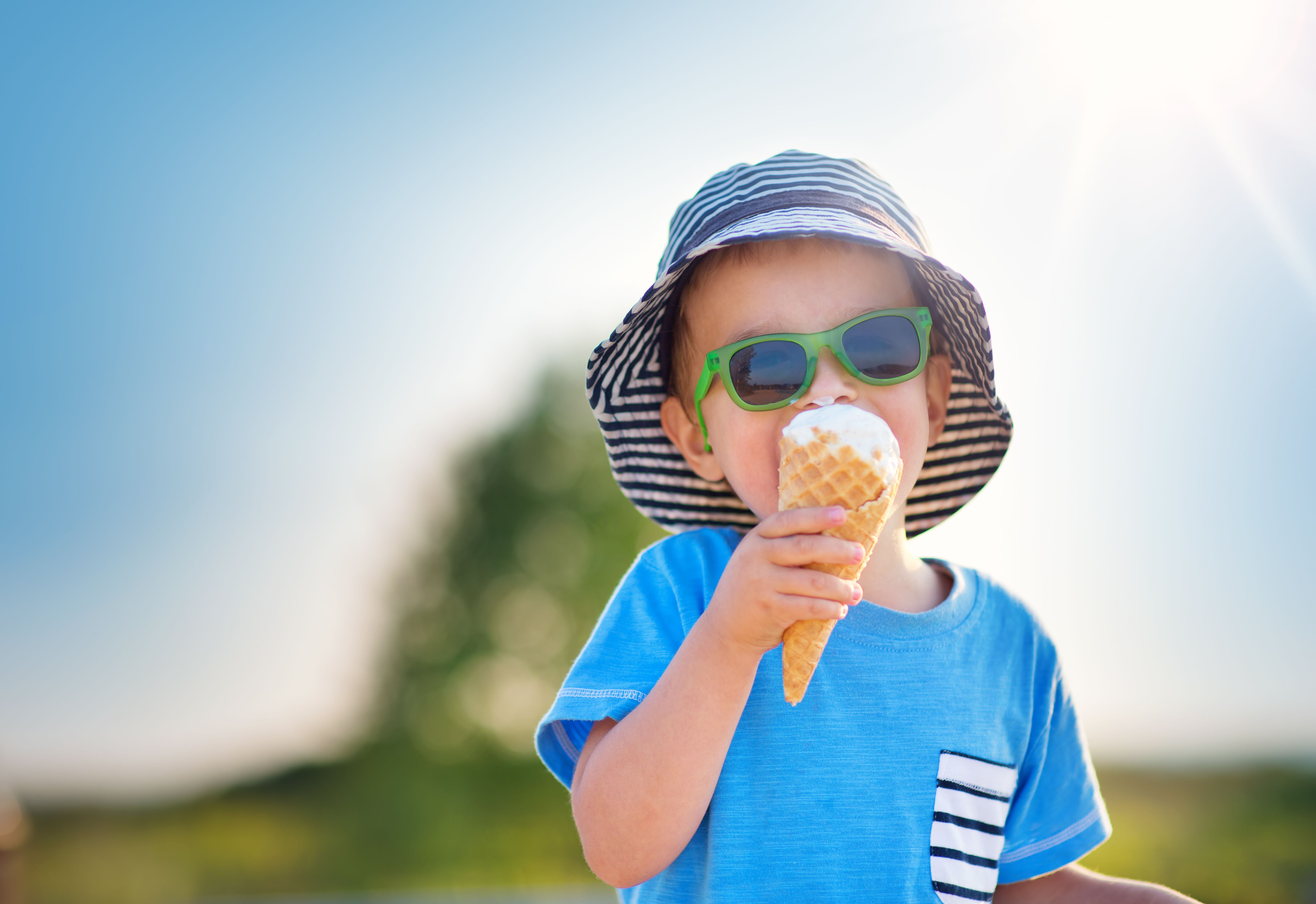 Junge mit Sonnenbrille isst im Sommer draußen ein Eis und trägt einen gestreiften Sonnenhut.