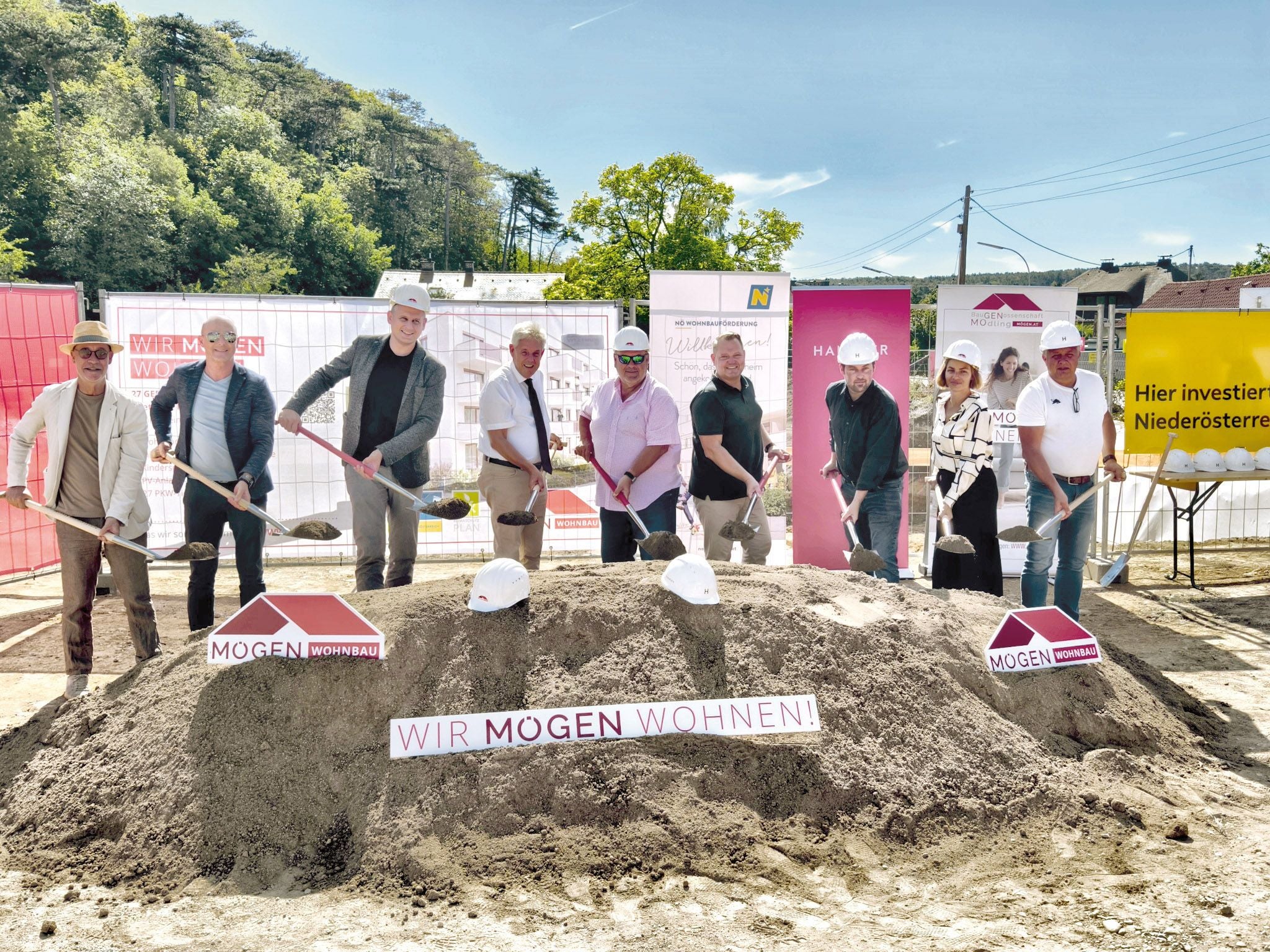 Mehrere Menschen mit Bauhelmen stehen bei einem offiziellen Spatenstich vor einem Sandhaufen.