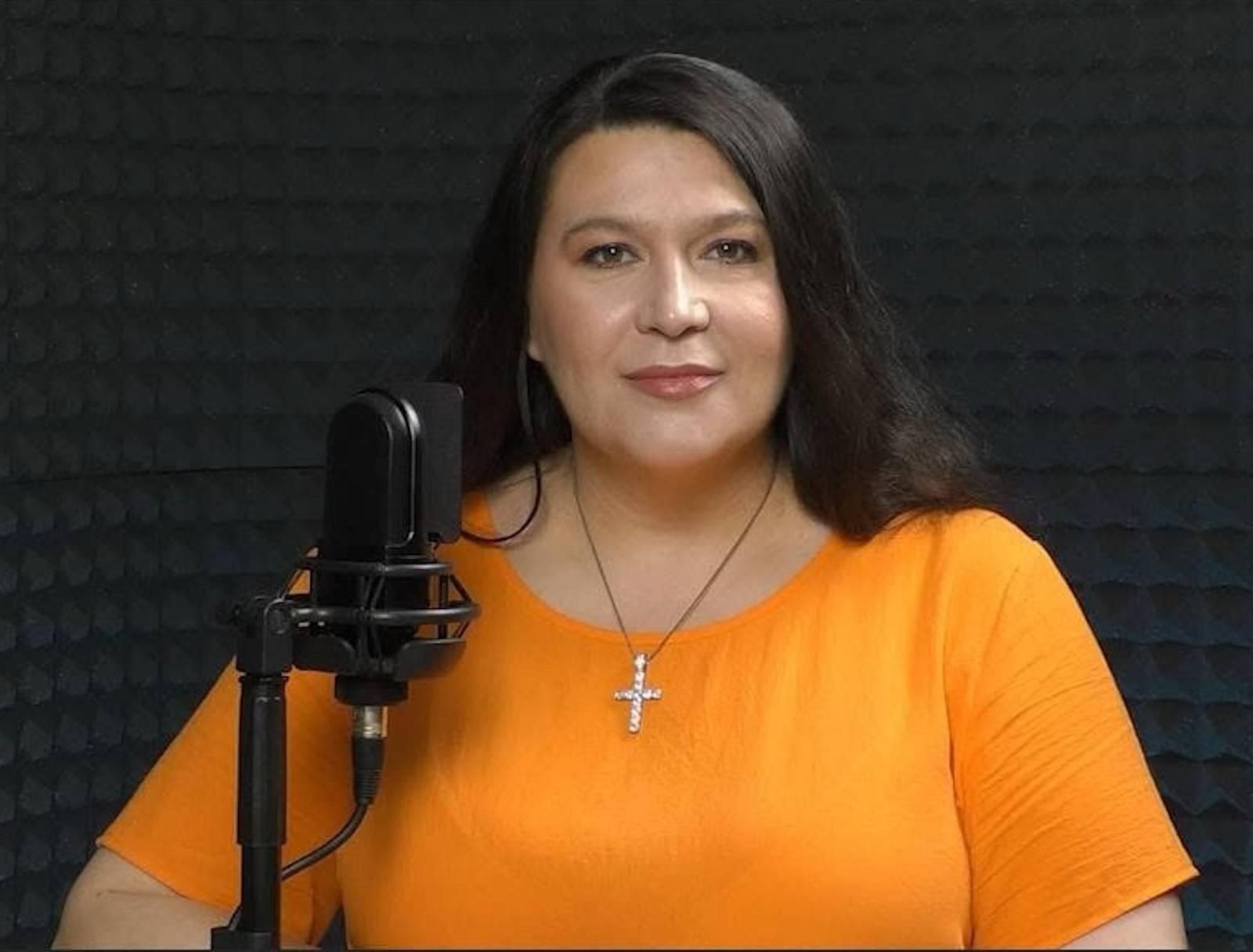 Frau im orangefarbenen Shirt sitzt vor einem Mikrofon in einem schallgedämmten Raum.