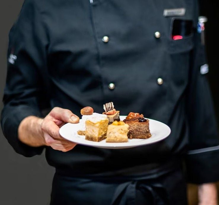 Koch in schwarzer Uniform hält einen Teller mit verschiedenen kleinen Kuchenstücken.
