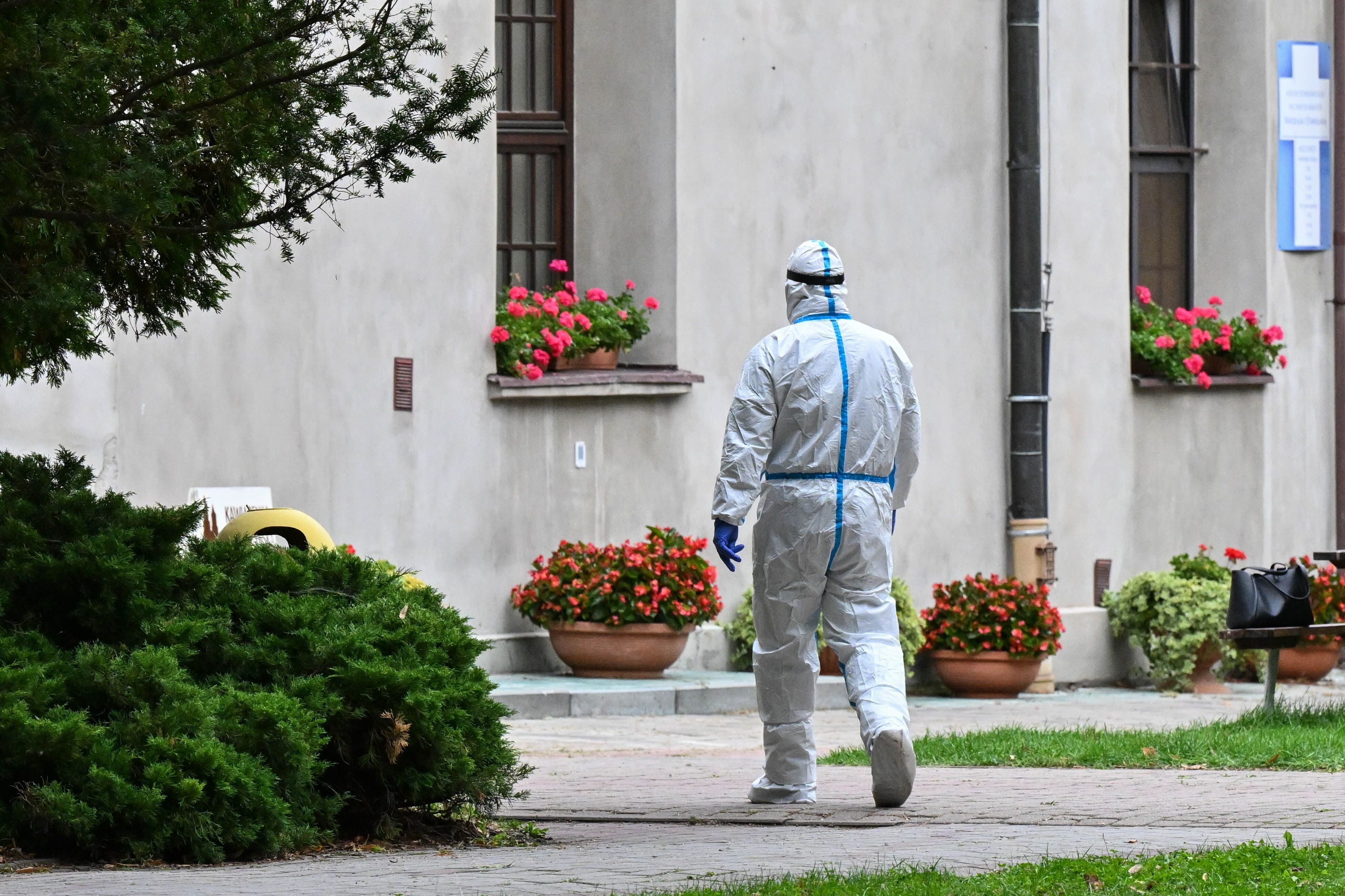 Eine Person in Schutzkleidung geht auf einem Gehweg vor einem Gebäude mit Blumenrabatten.