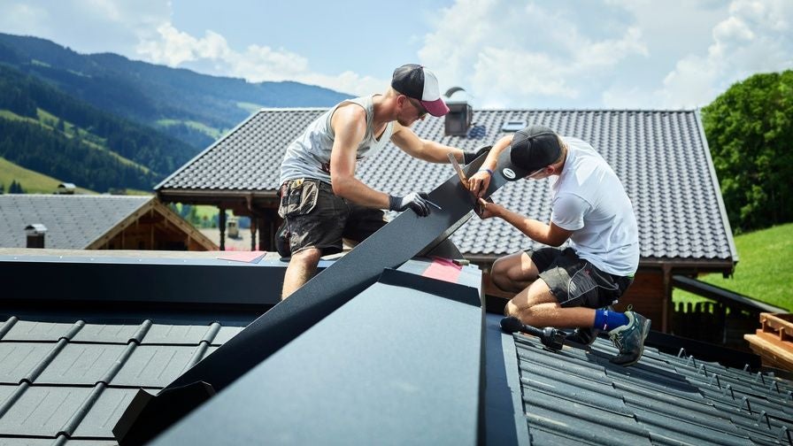 Zwei Handwerker arbeiten auf einem Dach vor alpiner Landschaft bei sonnigem Wetter.