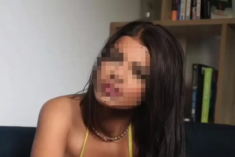 Frau mit langen dunklen Haaren sitzt vor einem Regal, ihr Gesicht ist verpixelt.