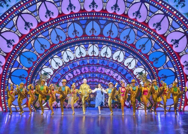 Bunte Musicalszene aus „Die Schöne und das Biest“ mit tanzendem Ensemble vor leuchtendem Bühnenbild.