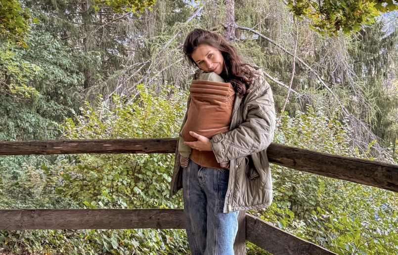 Eine Frau hält ein Baby in einer Trage auf einer Holzterrasse im Wald.