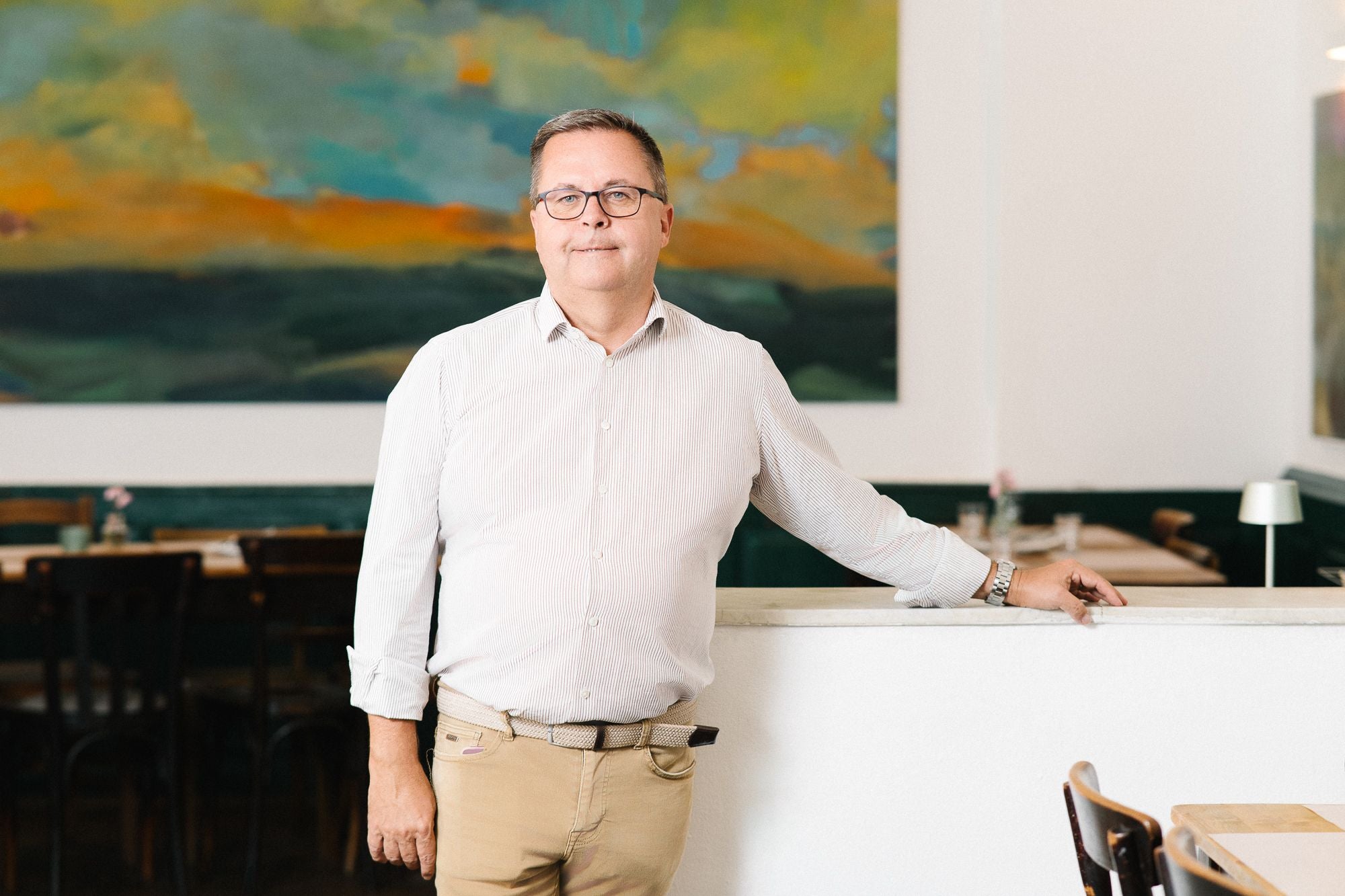 Ein Mann mit Brille steht in einem hellen Restaurant vor einem abstrakten Gemälde.