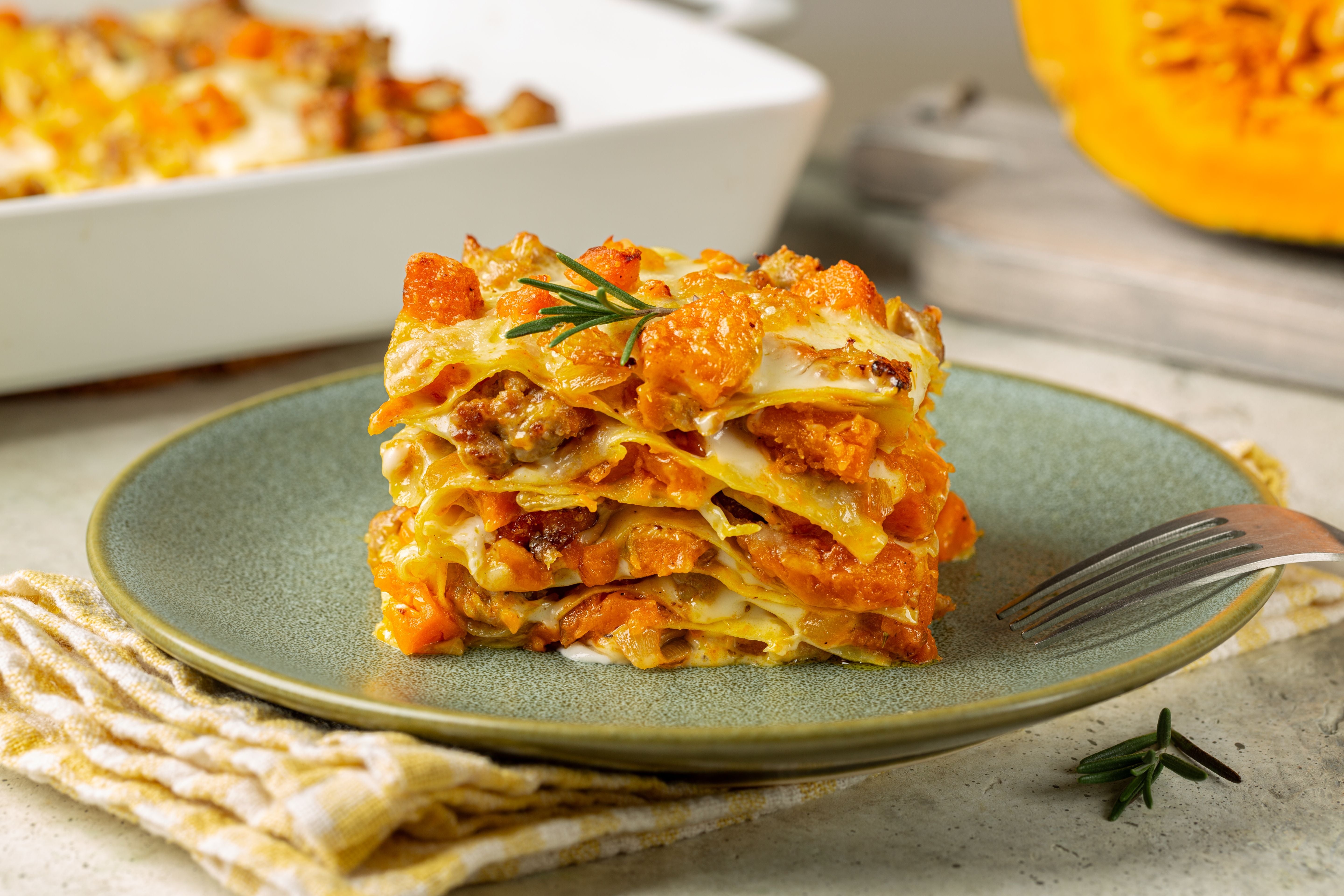Ein Stück Kürbis-Hackfleisch-Lasagne mit Käse auf einem grünen Teller, garniert mit Rosmarin.