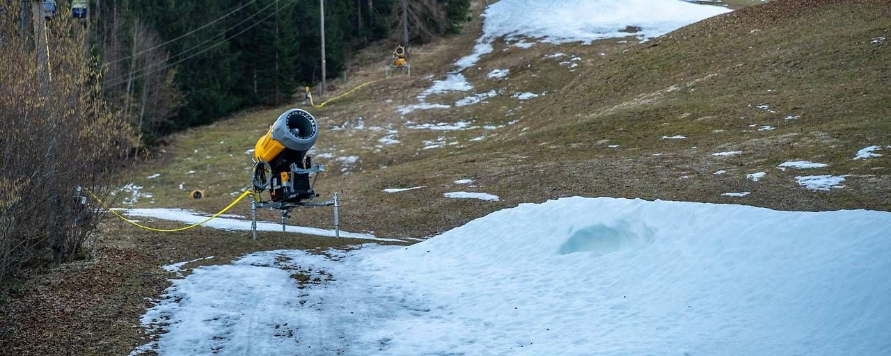 Eine Schneekanone steht auf einer schneearmen Piste mit vereinzelten Schneeflecken.