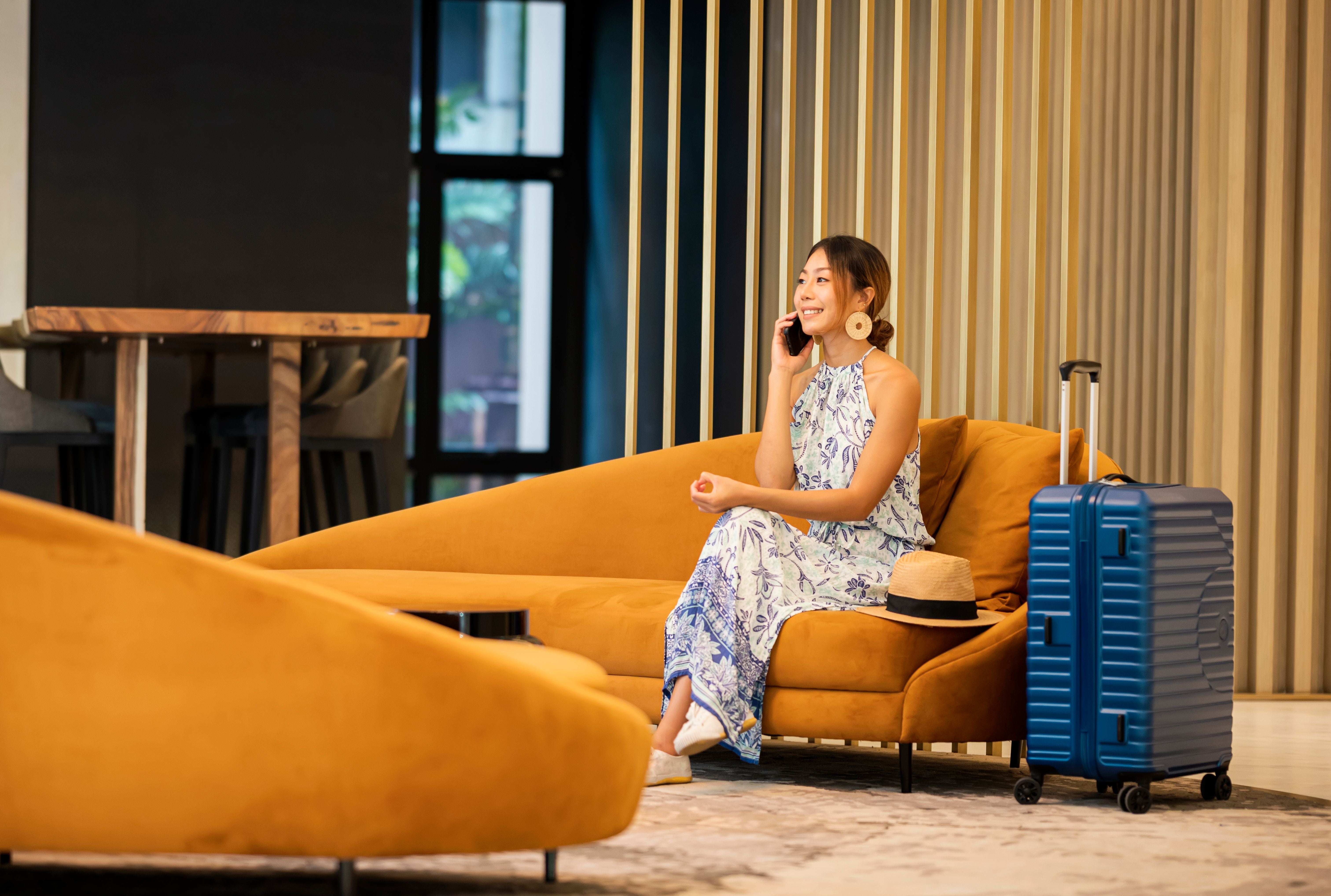 Junge Frau sitzt in Hotellounge auf Sofa, telefoniert und hat blauen Koffer neben sich.