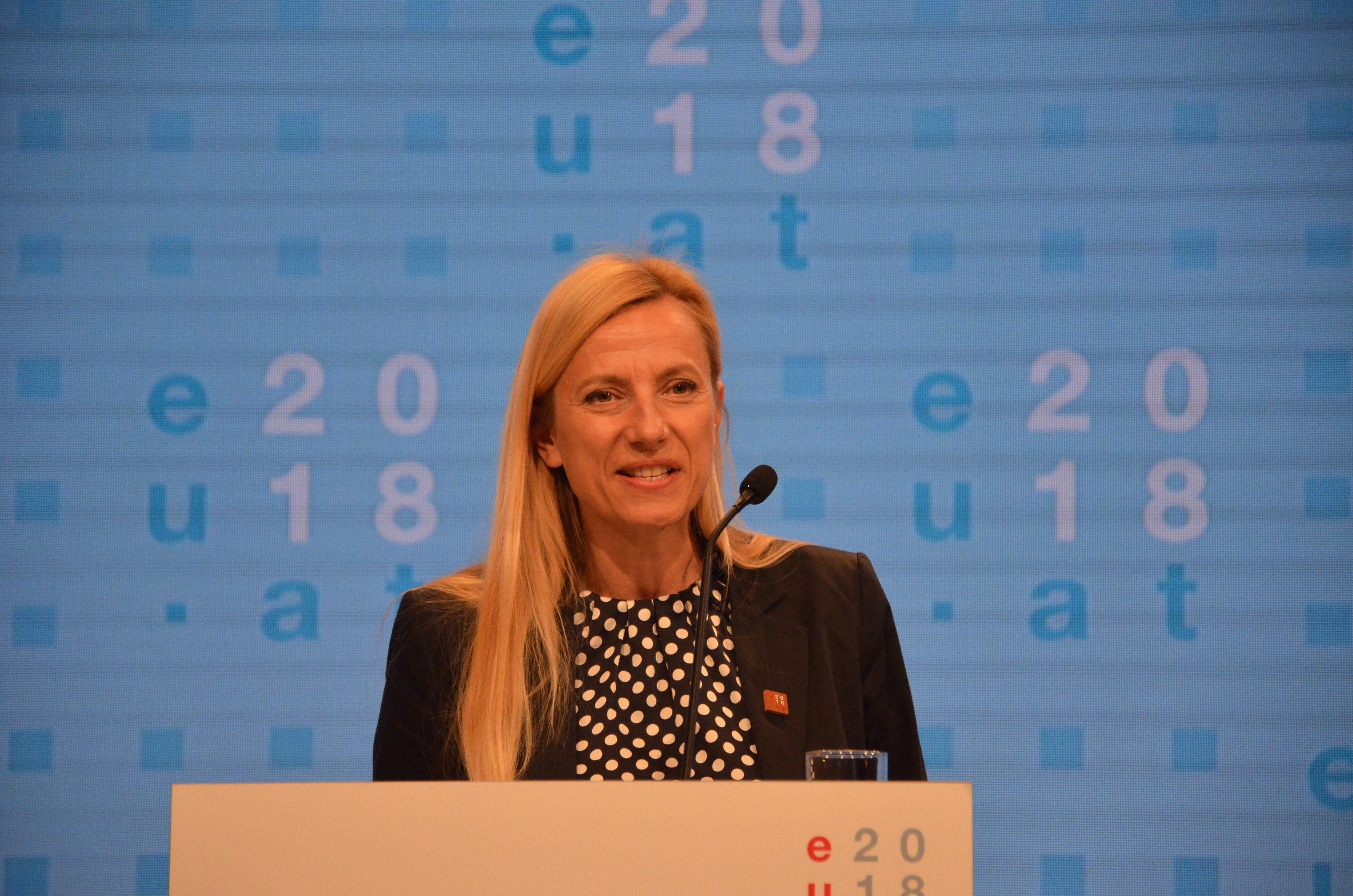 Frau am Rednerpult bei einer Konferenz mit blauem Hintergrund und Mikrofon.