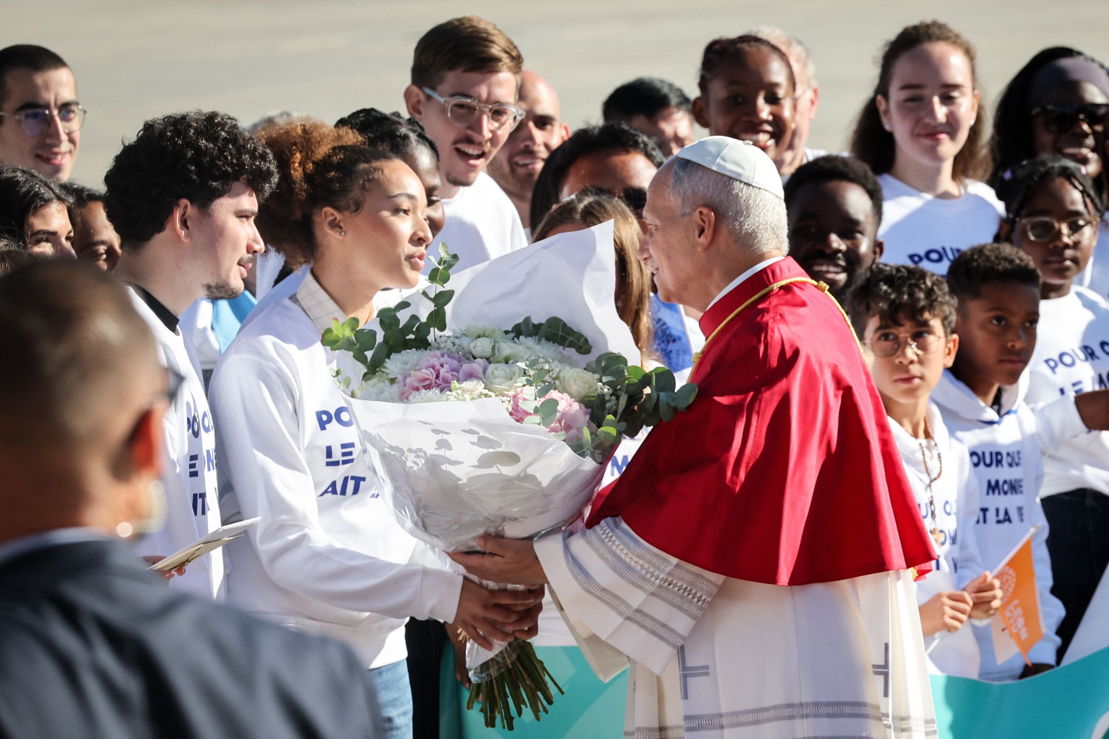 Papst in rotem Gewand erhält am Flughafen einen Blumenstrauß von einer jungen Frau, umgeben von Menschen.