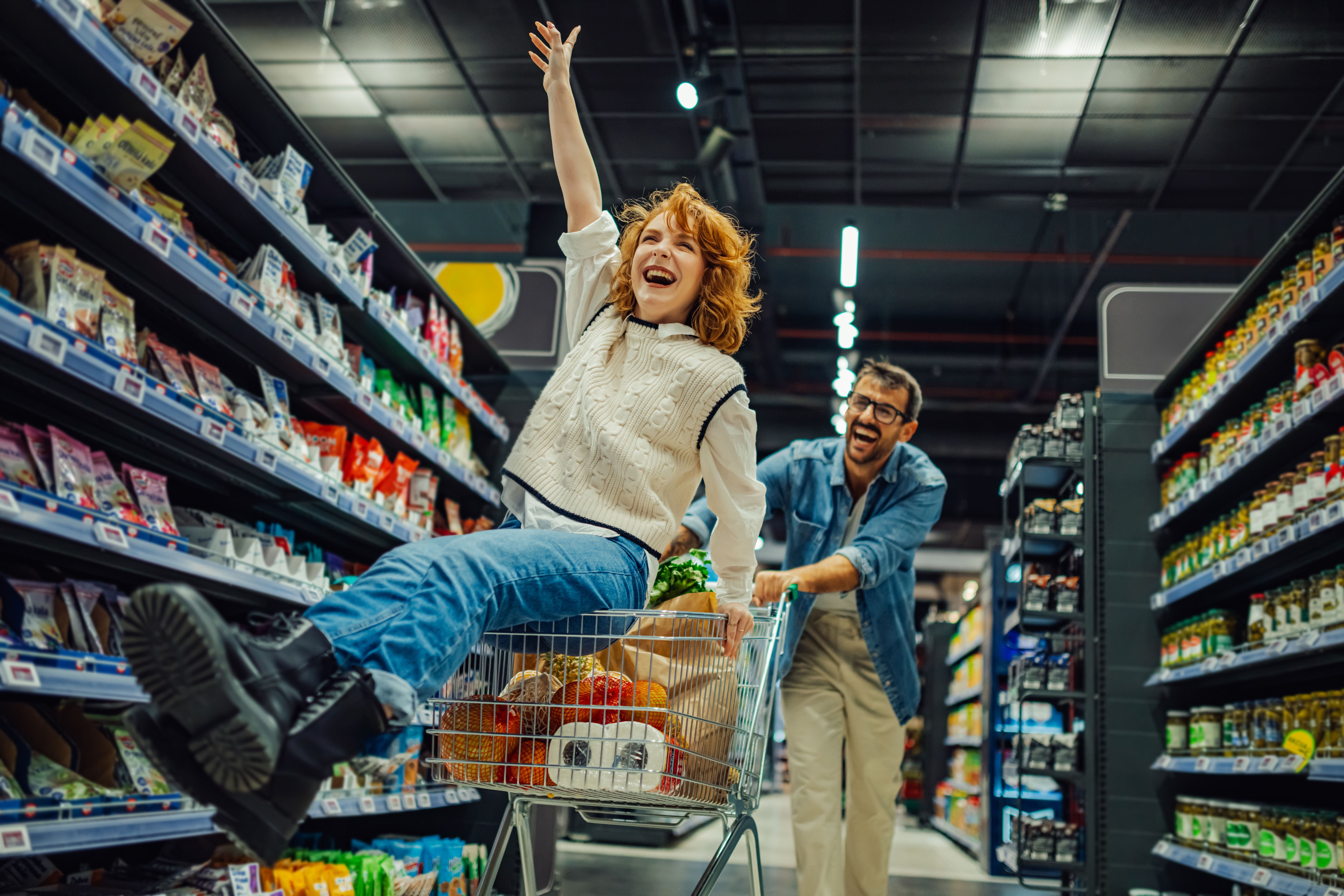 Junge Frau lacht und sitzt im Einkaufswagen, der von einem Mann durch den Supermarkt geschoben wird.