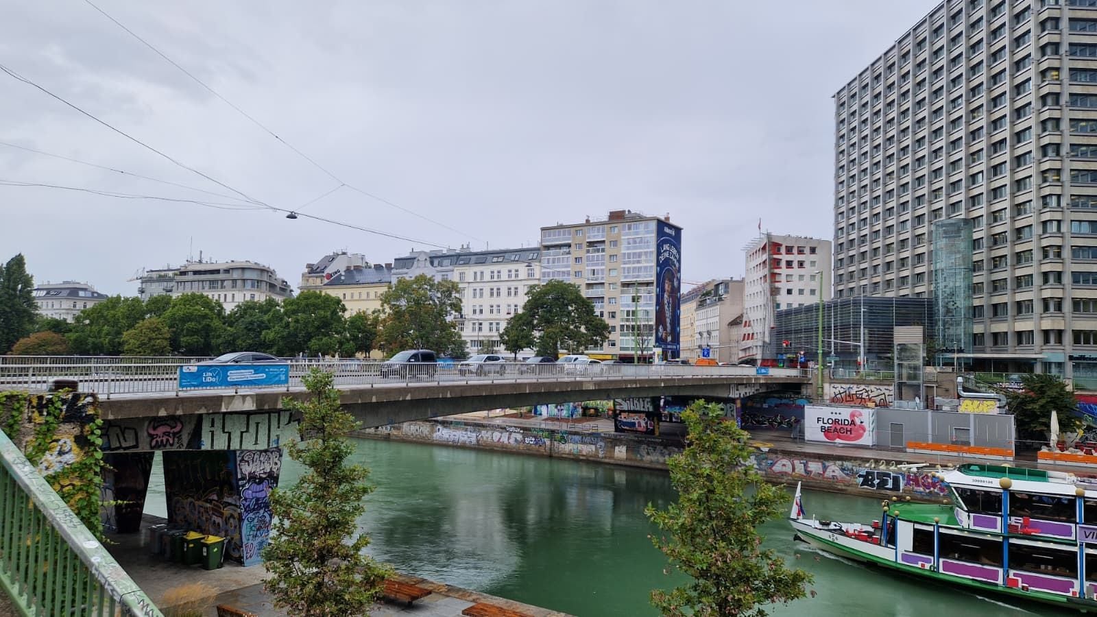 Die Salztorbrücke in Wien überspannt den Donaukanal, Autos fahren, ein Ausflugsschiff fährt darunter durch.