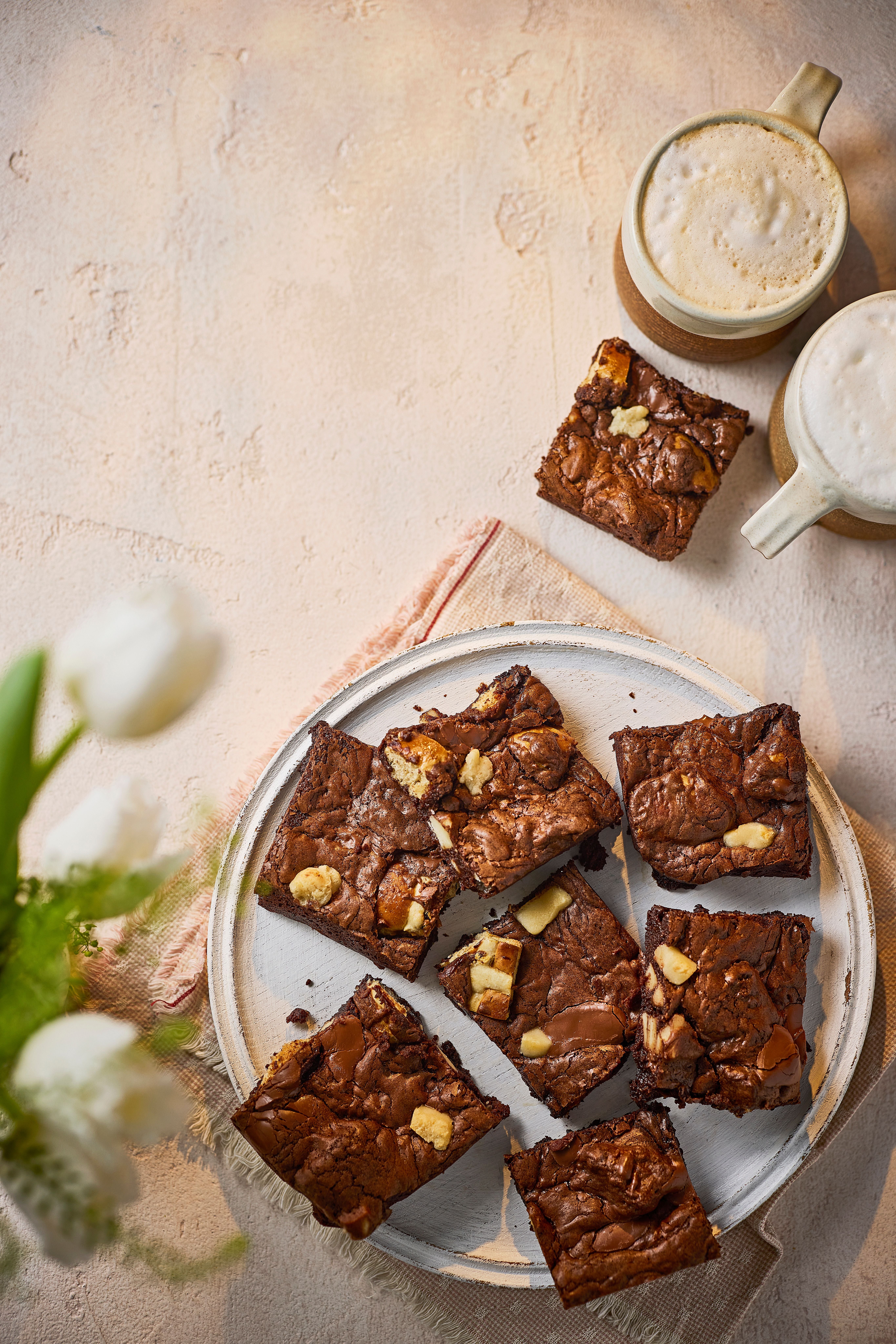 Mehrere Brownies mit Stückchen und zwei Tassen Kaffee auf einem hellen Tisch.