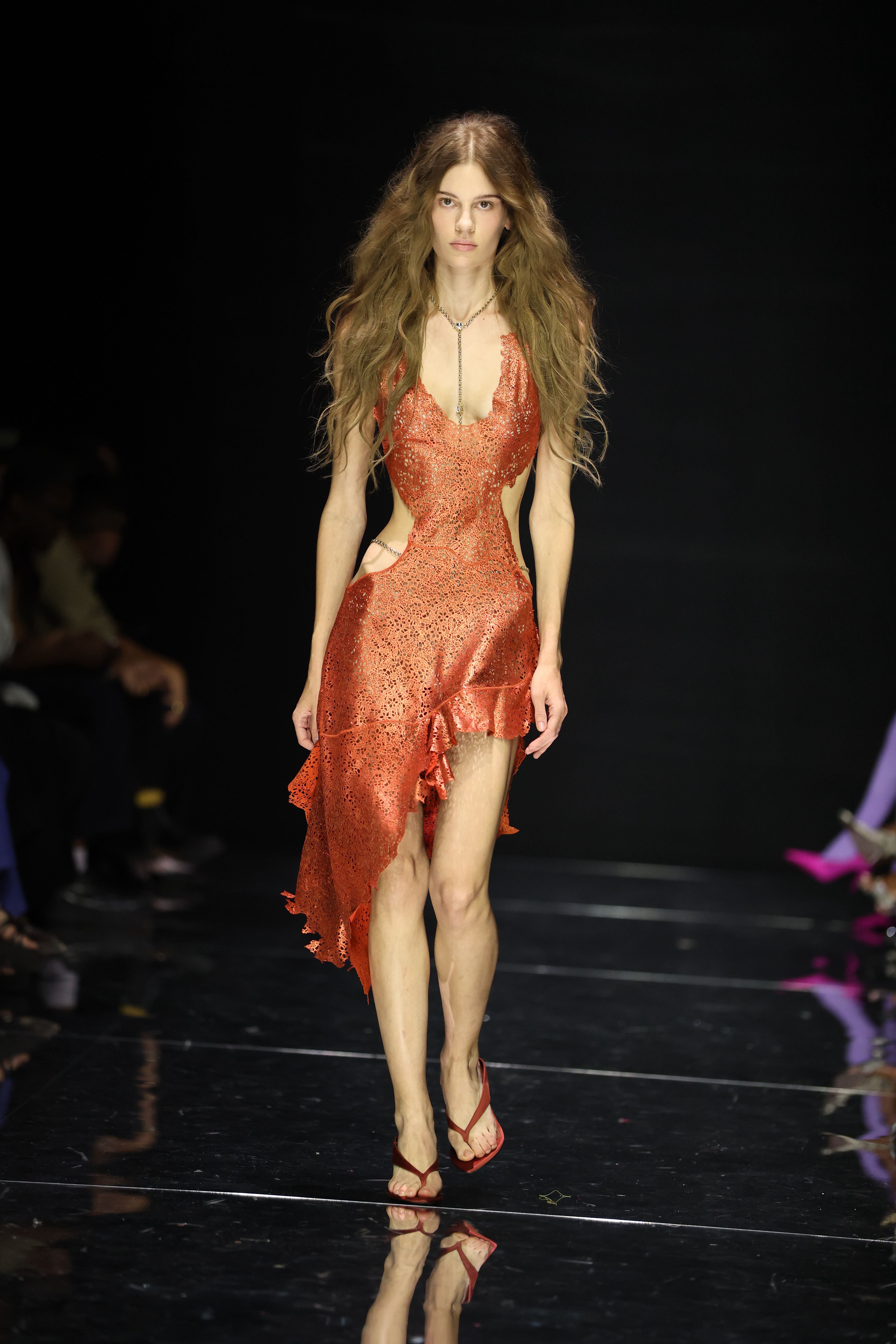 Model in asymmetrischem, orangefarbenem Kleid auf dem Laufsteg bei einer Modenschau.
