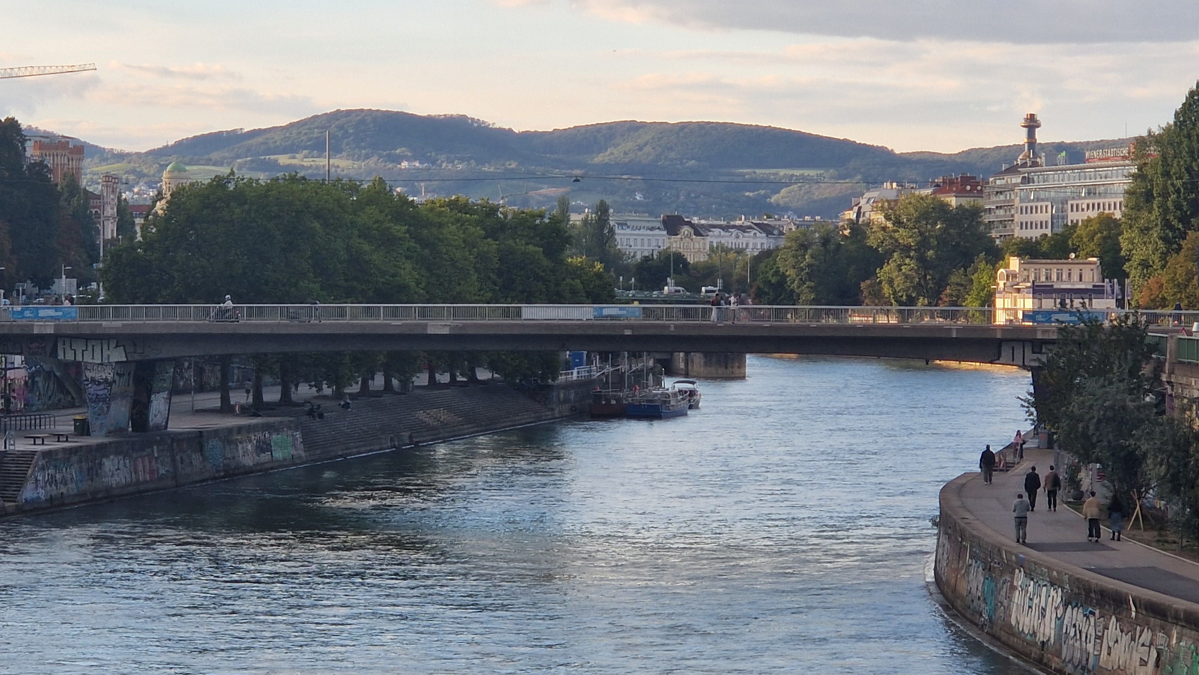 Brücke über den Donaukanal in Wien mit Spaziergängern am Ufer und Hügeln im Hintergrund.