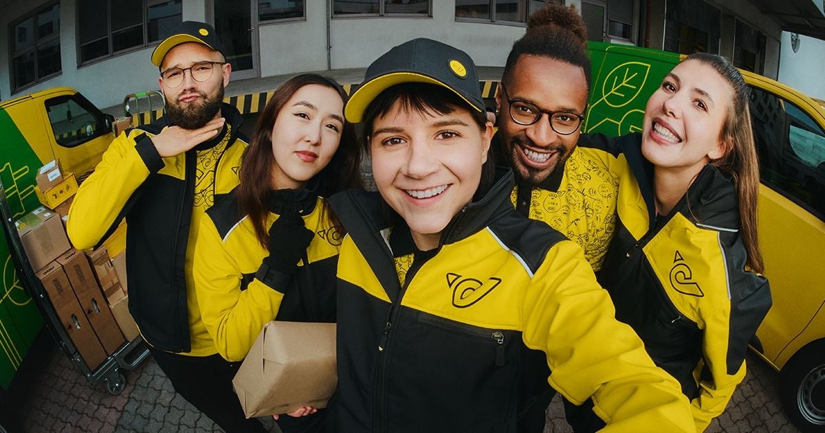 Fünf Paketzusteller in gelber Uniform posieren lachend vor einem Lieferwagen.