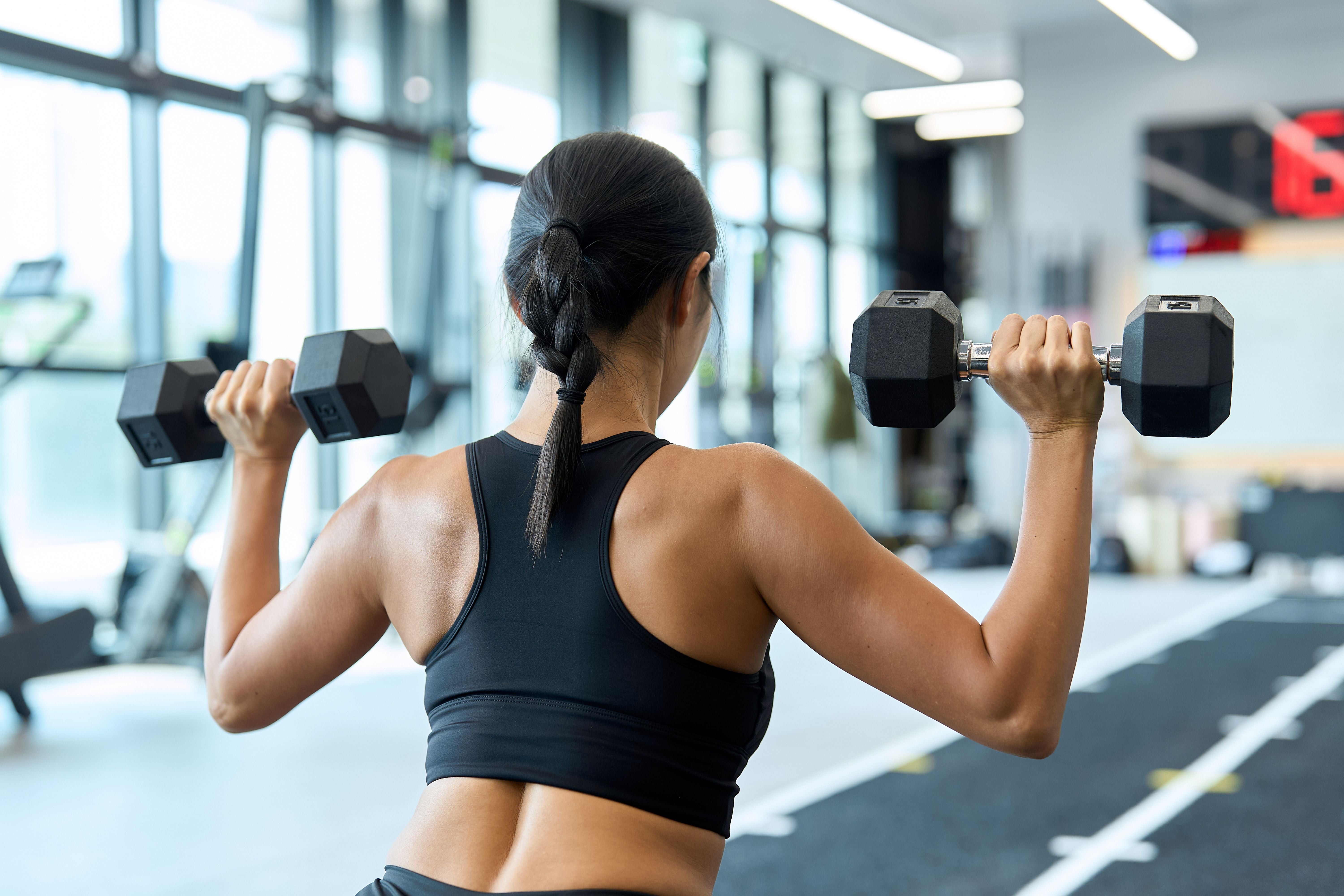 Frau im Fitnessstudio hebt zwei Kurzhanteln, Blick von hinten auf Rücken und Arme.
