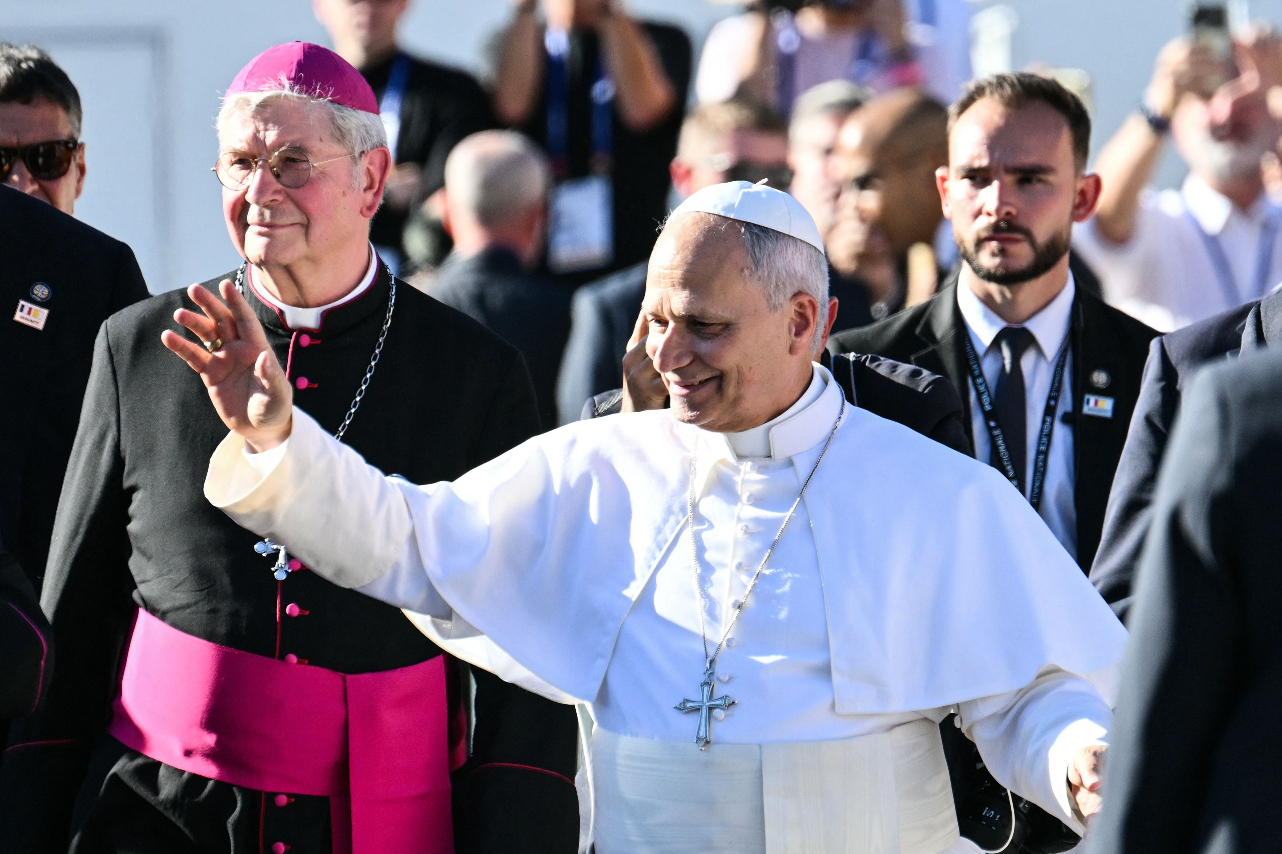 Papst in weißer Kutte winkt, begleitet von Bischof und Sicherheitsleuten, Menschenmenge im Hintergrund.
