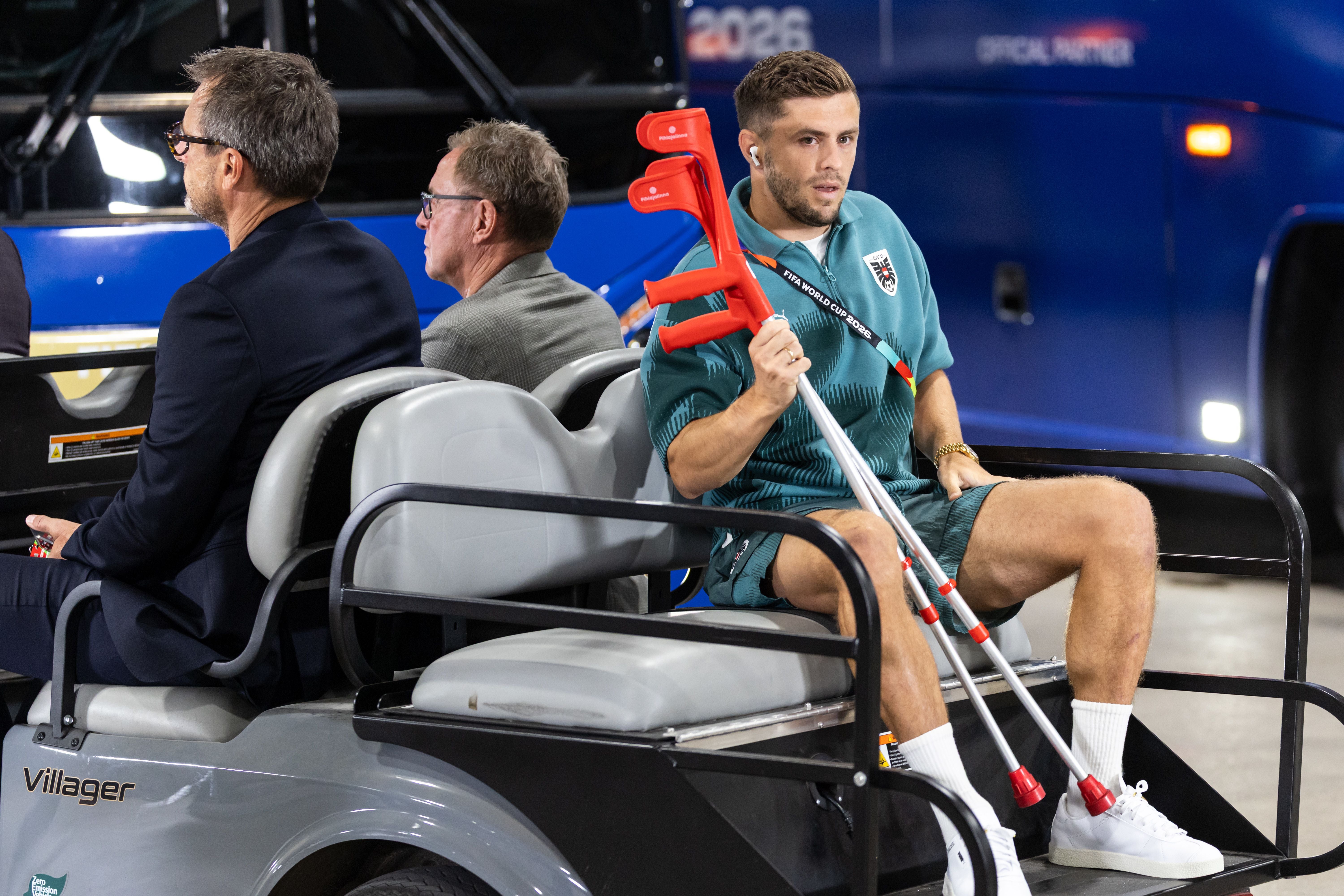 Ein verletzter Fußballspieler mit Krücken sitzt mit zwei Männern auf einem Golfcart.