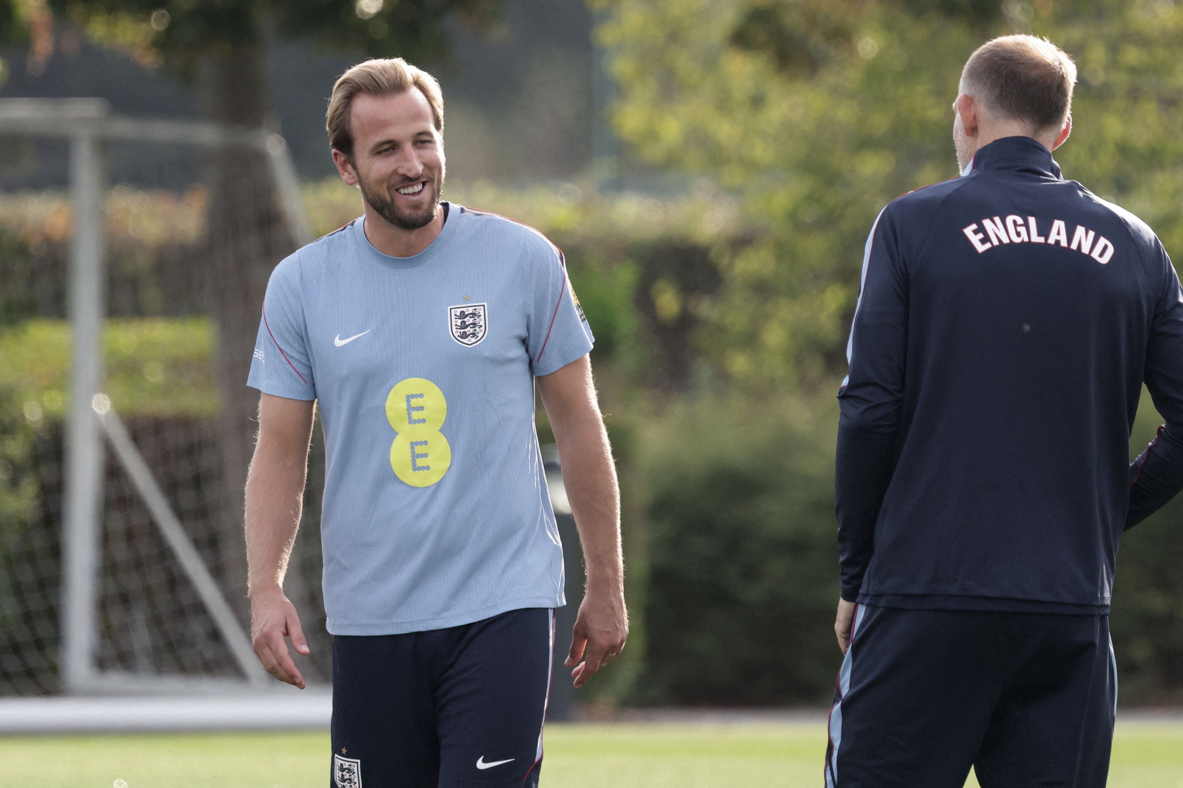 Harry Kane lächelt im hellblauen Trainingsshirt der englischen Nationalmannschaft neben Trainer Thomas Tuchel von hinten.