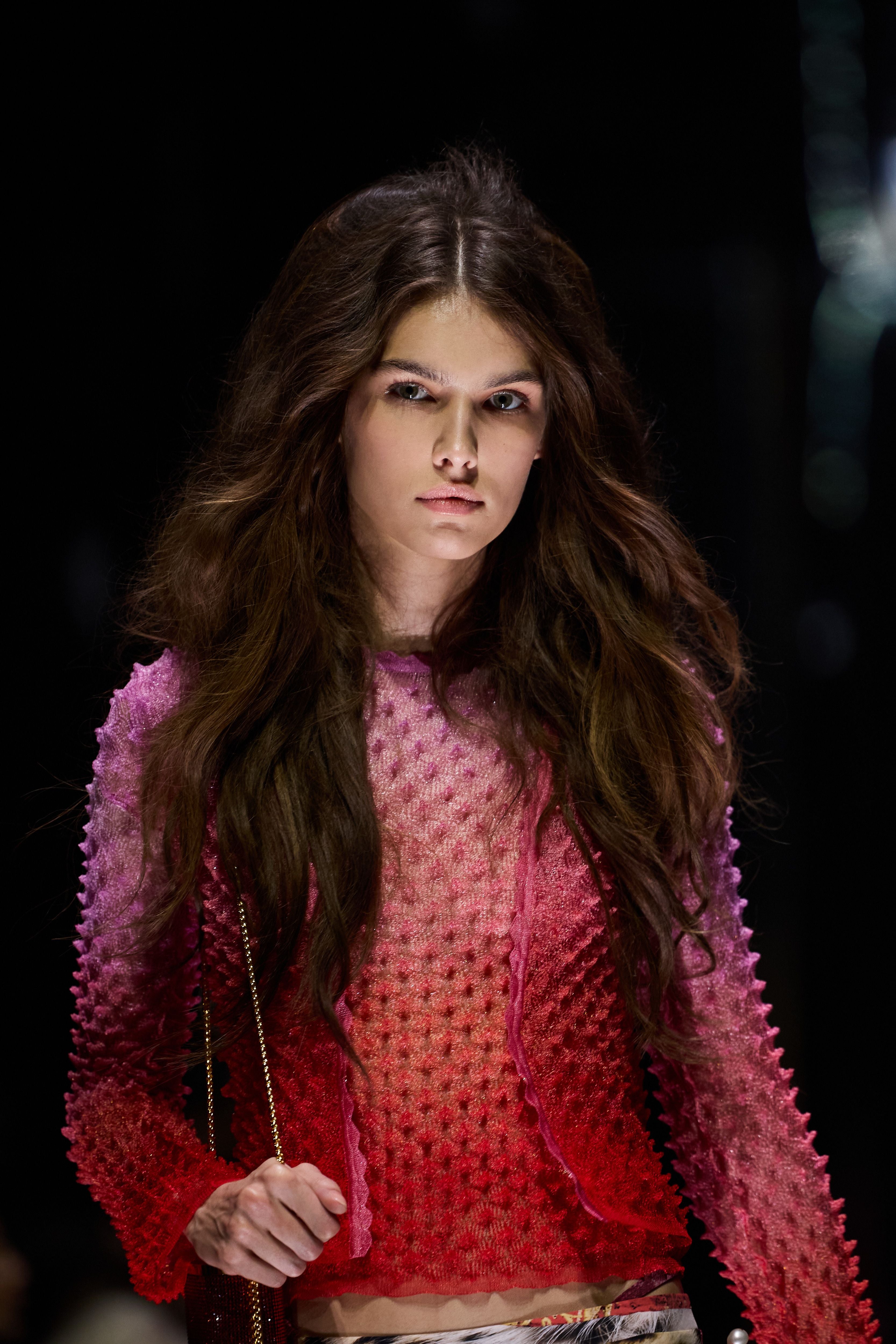 Model mit langen braunen Haaren trägt ein rosa-rotes Oberteil auf dem Laufsteg.