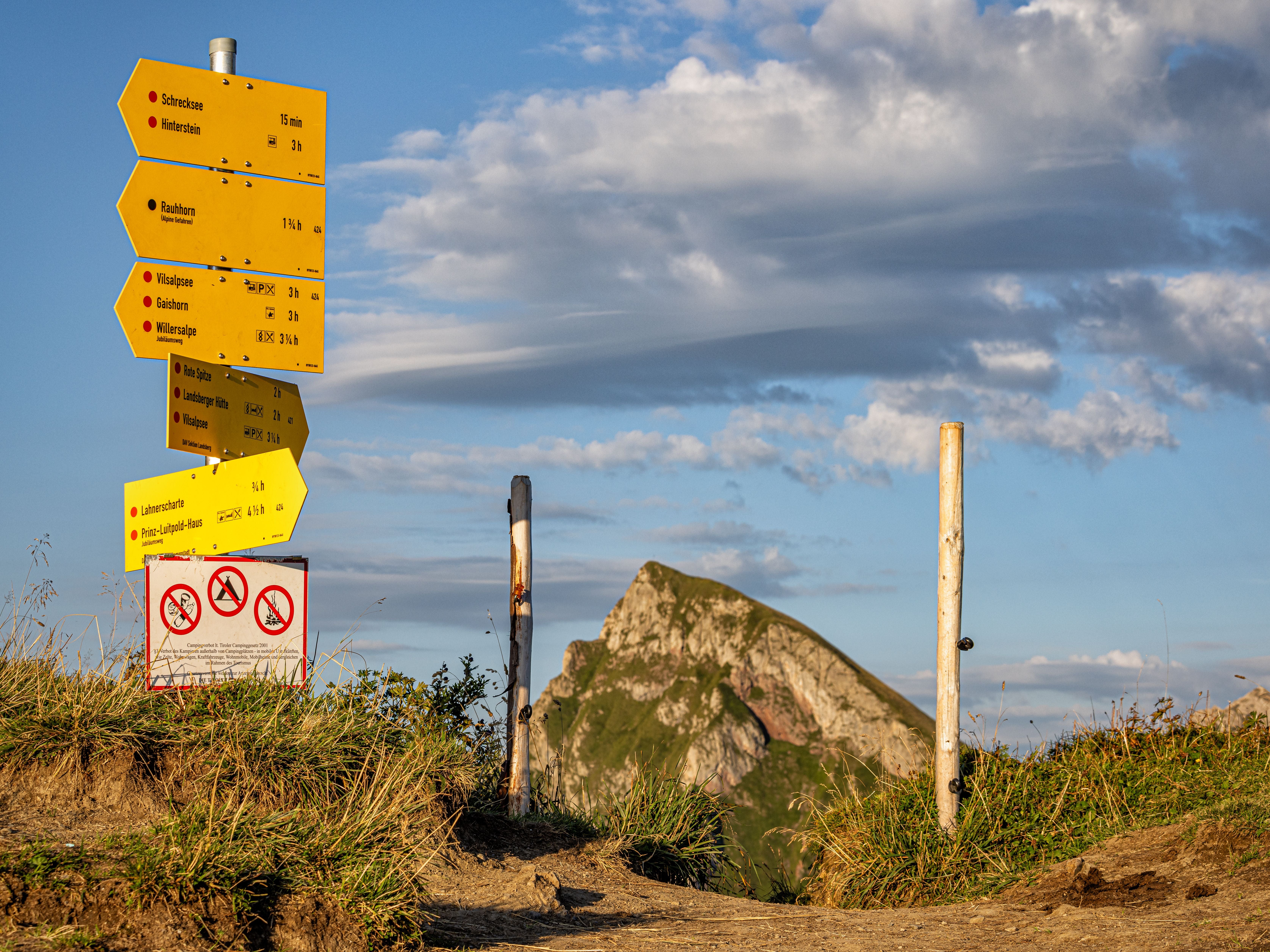 Gelbe Wanderschilder und Verbotsschilder auf einem Bergpfad mit Gipfel im Hintergrund.
