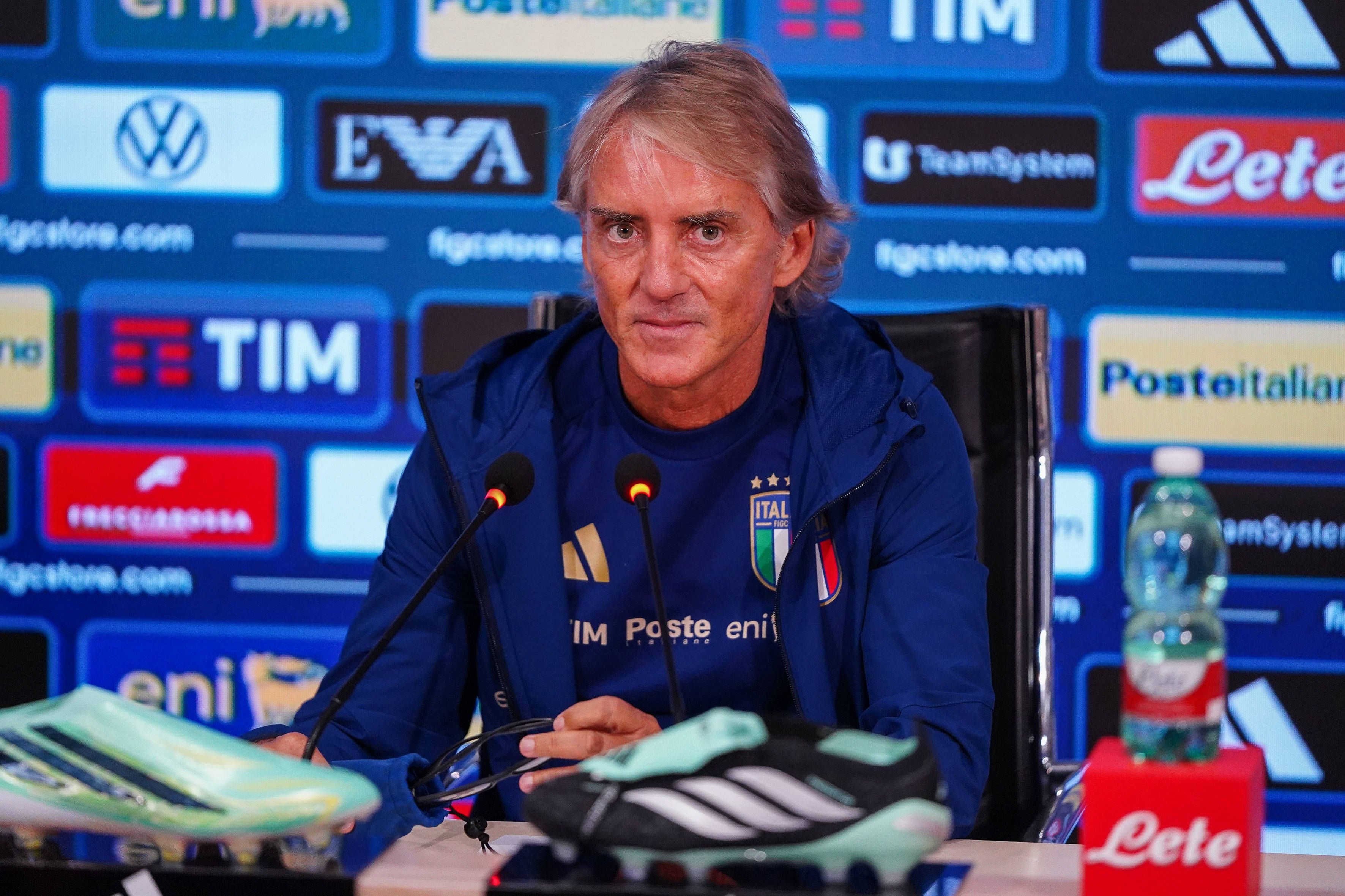 Roberto Mancini sitzt im blauen Trainingsanzug der italienischen Nationalmannschaft bei einer Pressekonferenz hinter Mikrofonen.