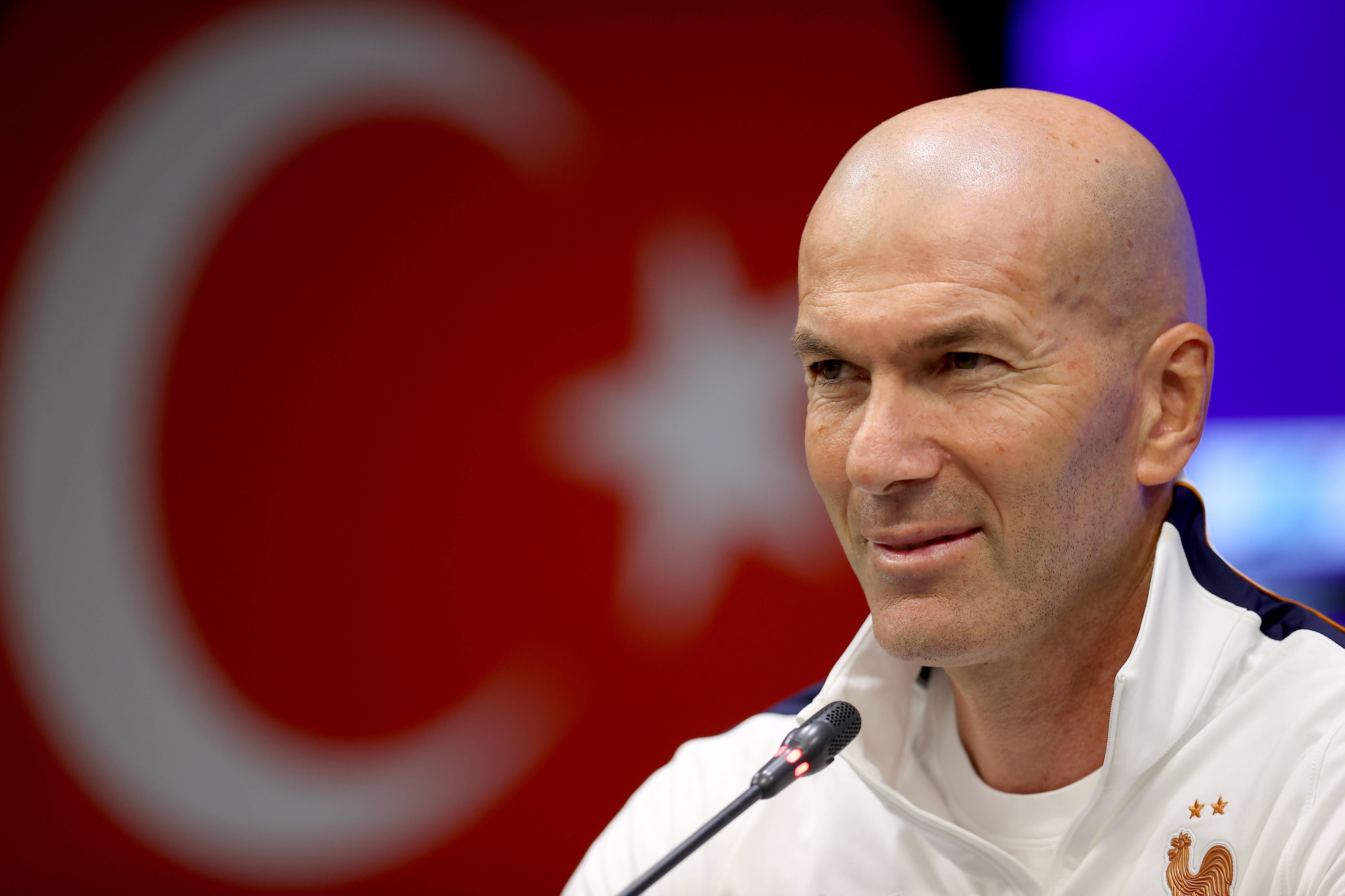 Zinedine Zidane blickt bei der Pressekonferenz im Trainingsanzug der französischen Nationalmannschaft lächelnd zur Seite.