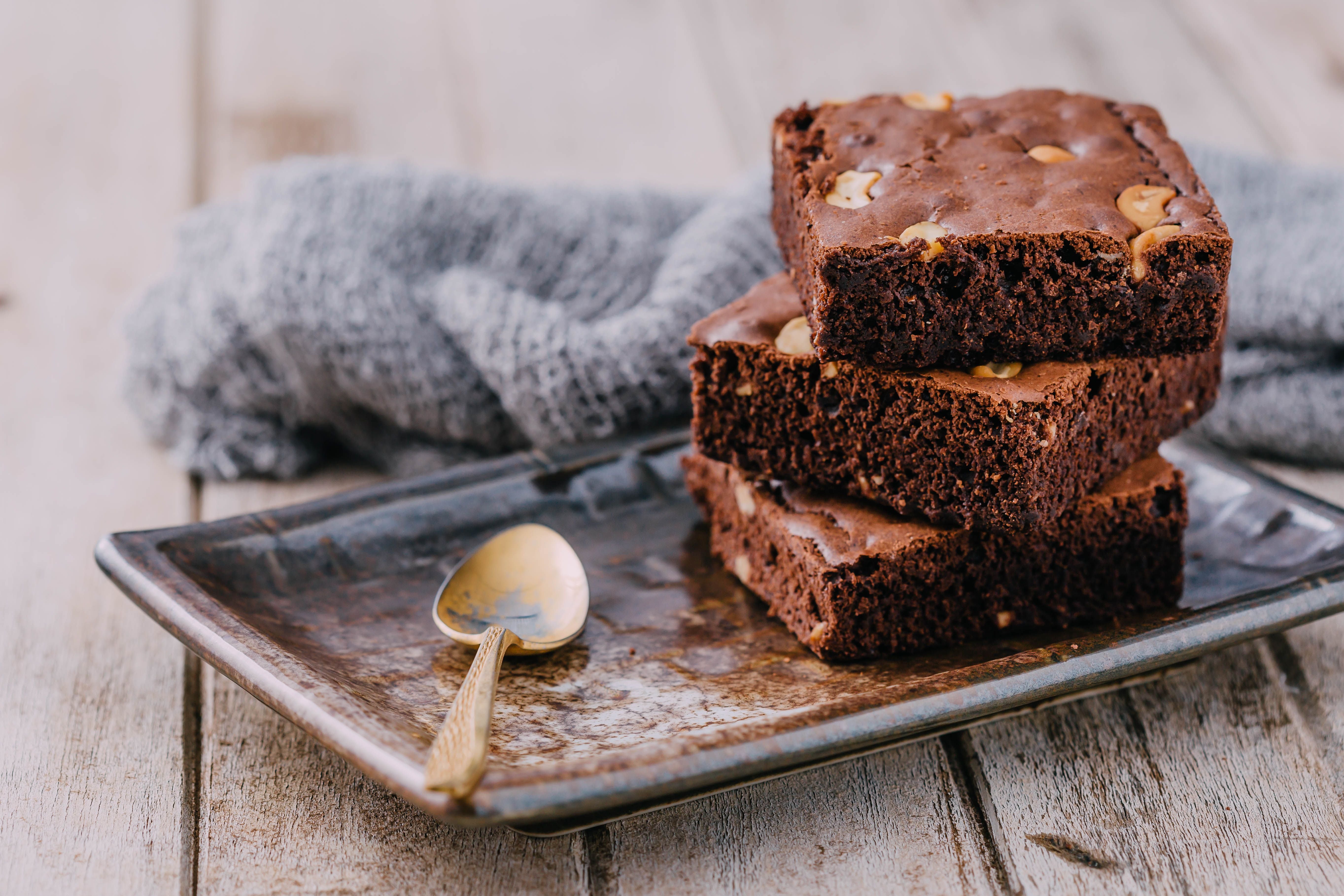 Drei Brownie-Stücke liegen gestapelt auf einem Blech neben einem Löffel und grauem Tuch.