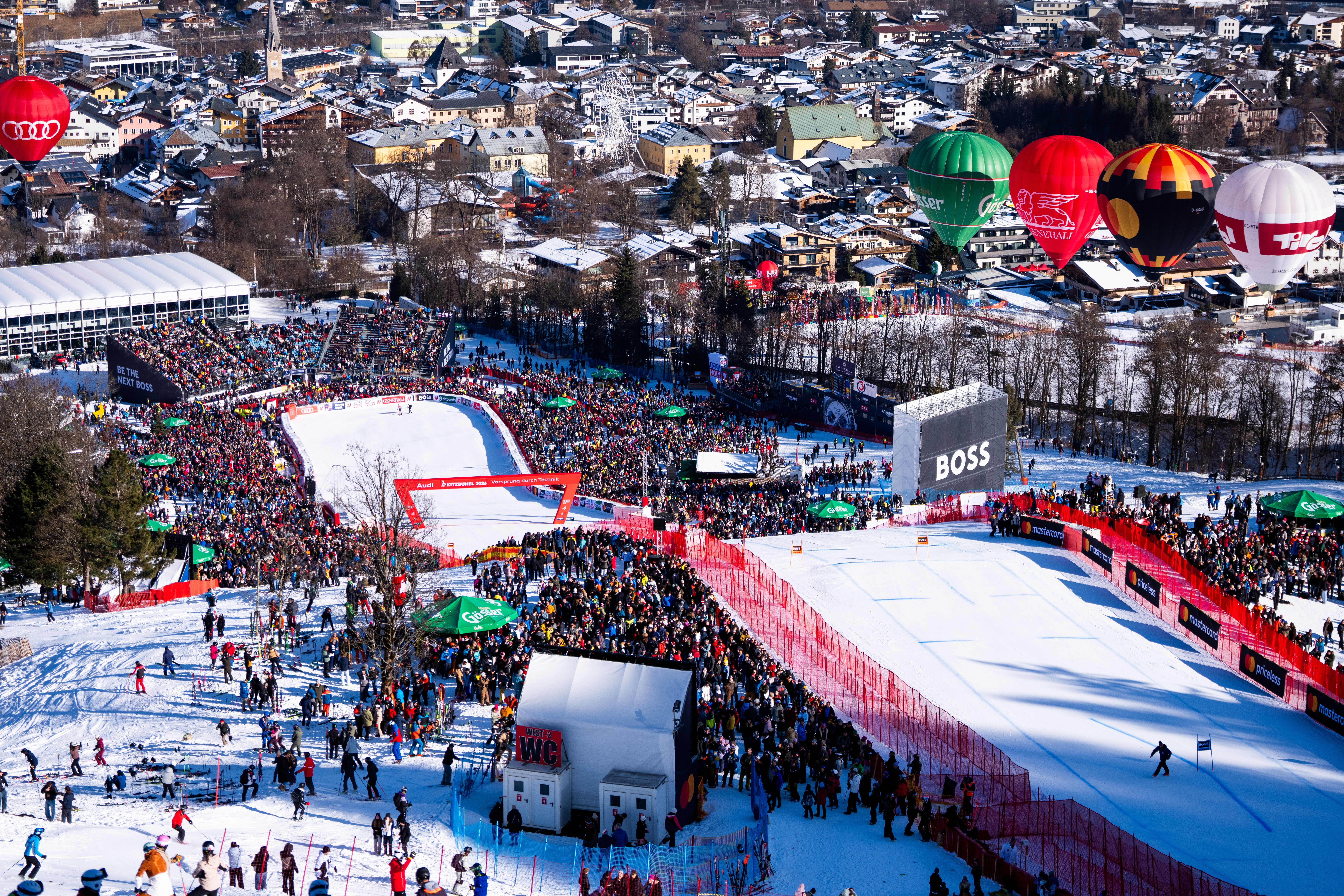Viele Zuschauer verfolgen ein Ski-Skirennen im Zielbereich, im Hintergrund bunte Heißluftballons.
