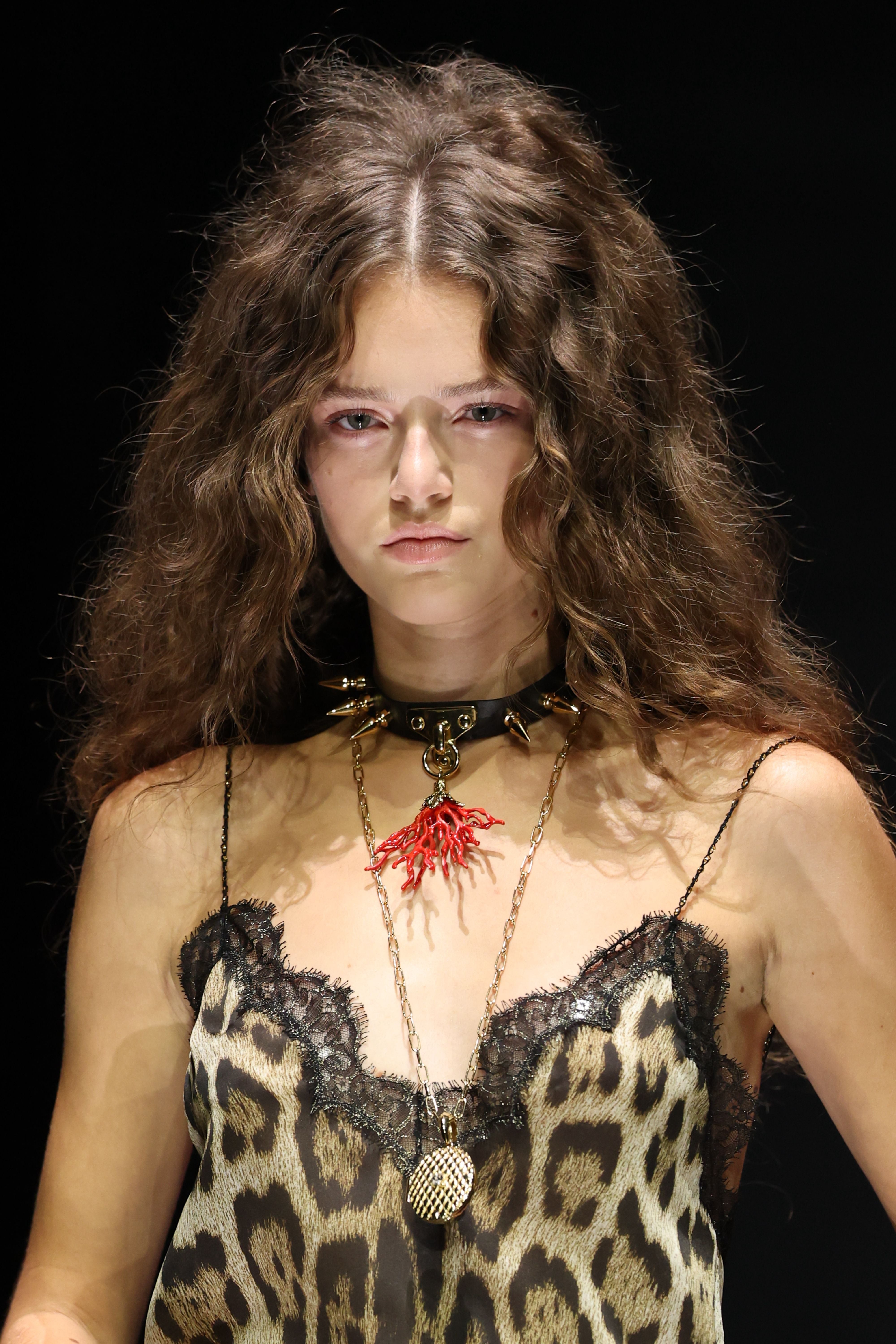 Model mit langen, lockigen Haaren trägt Leopardenkleid und auffällige Ketten auf dem Laufsteg.