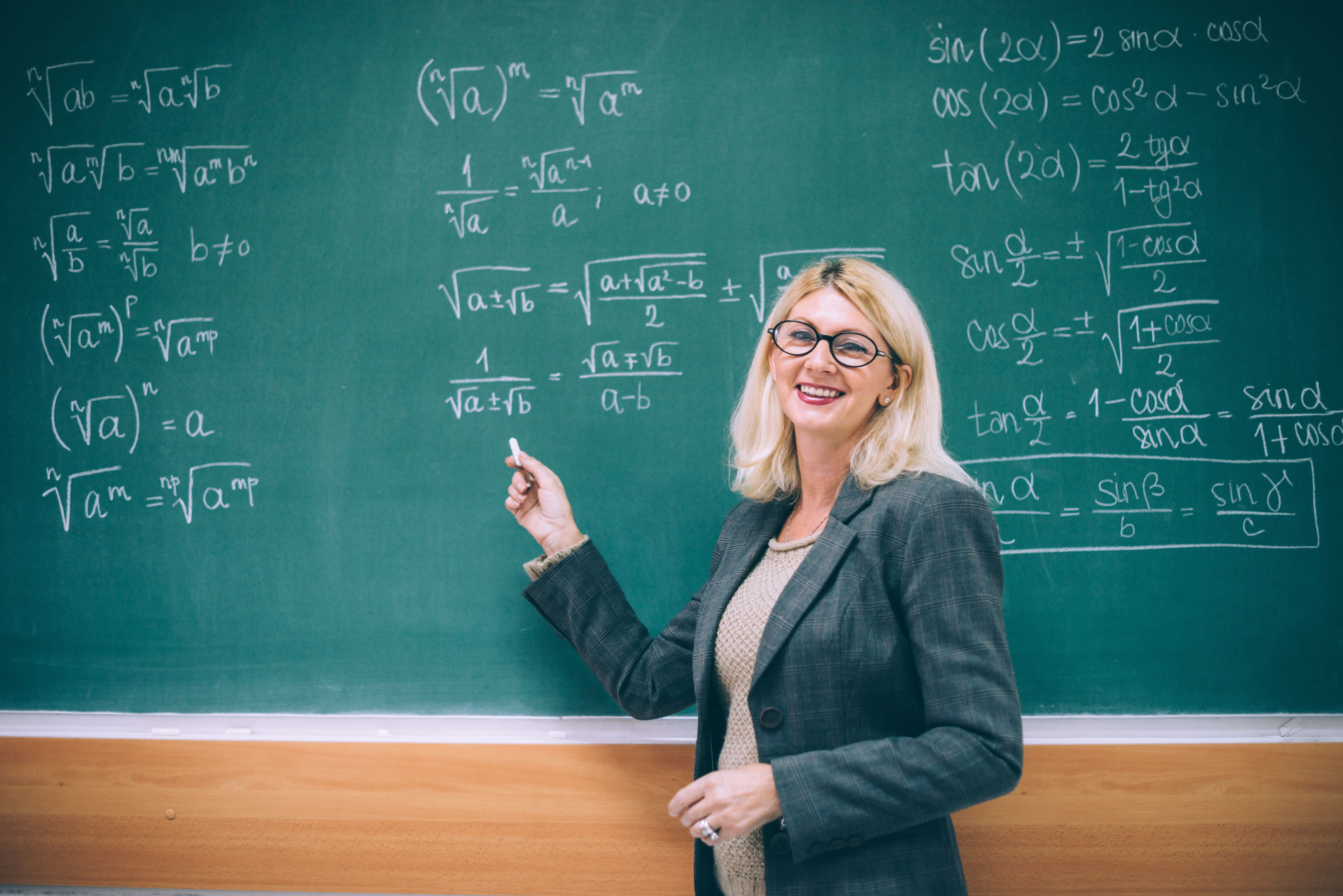Lehrerin steht lächelnd vor einer Tafel mit mathematischen Formeln und hält Kreide in der Hand.