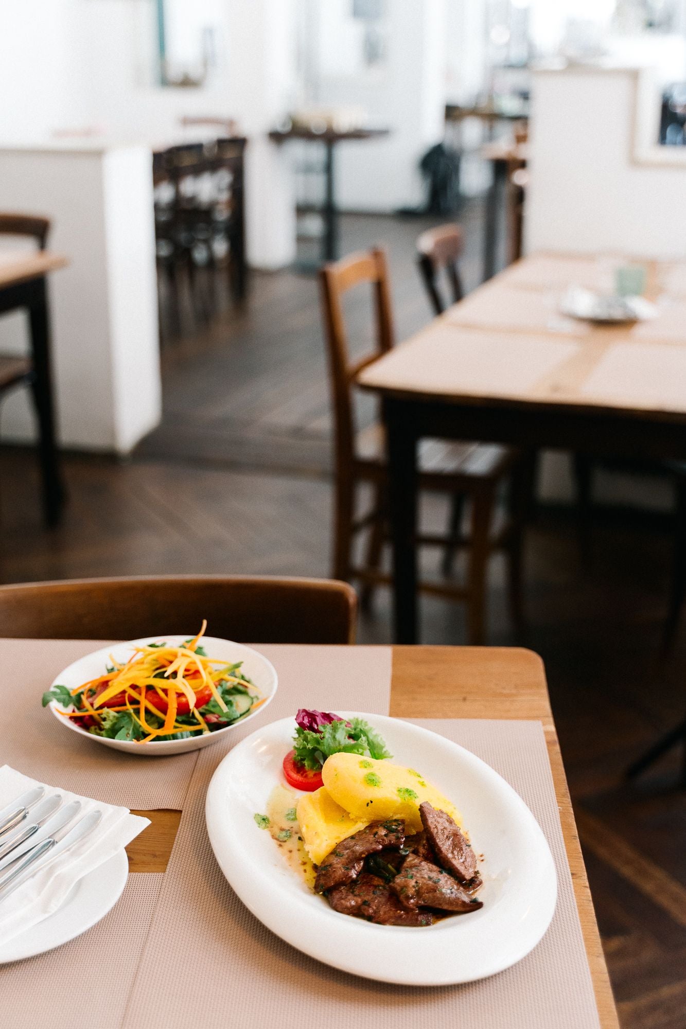 Ein gedeckter Restauranttisch mit Salat und Fleischgericht mit Kartoffeln auf weißen Tellern.