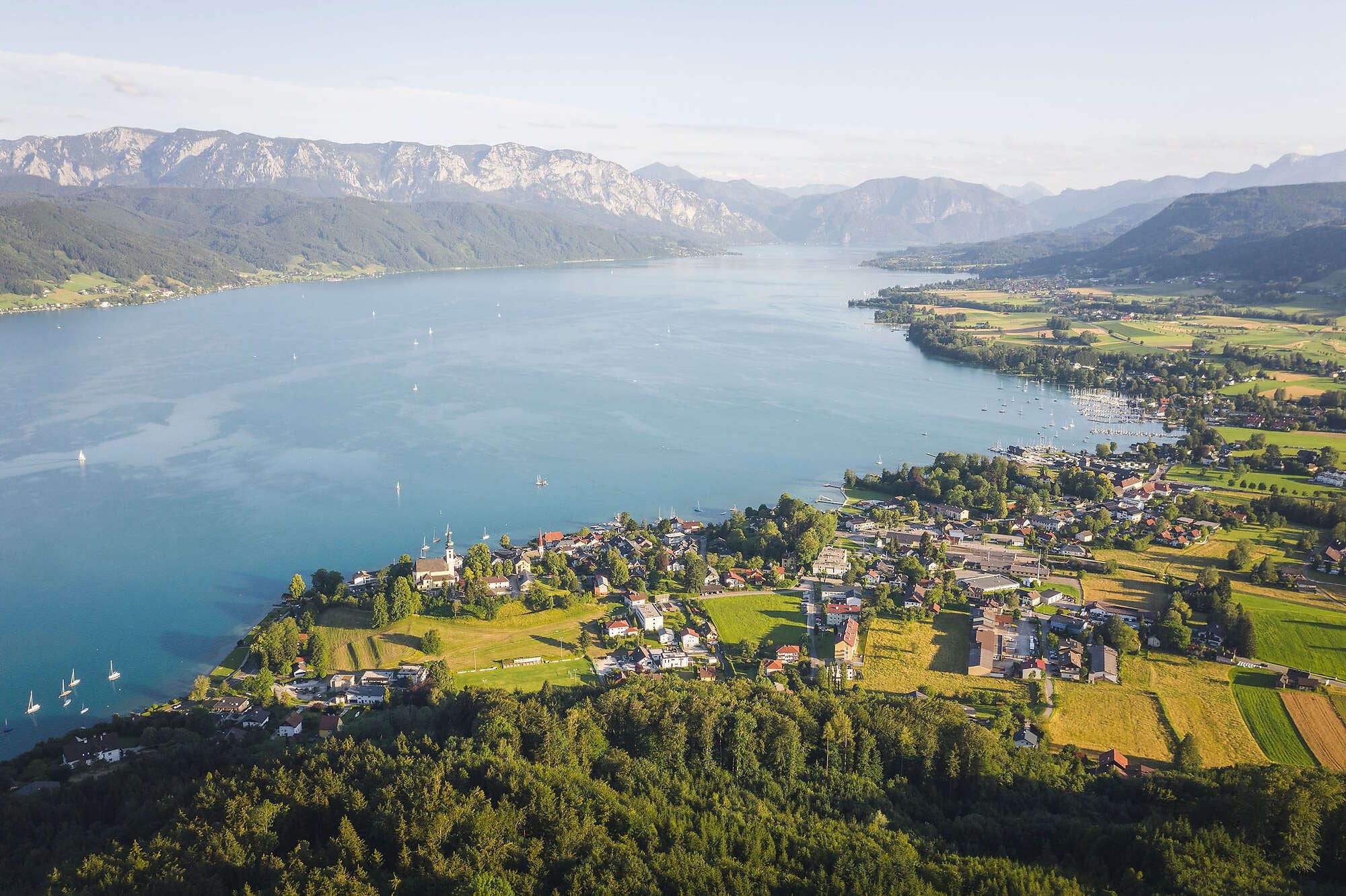 Luftaufnahme des Attersees mit umliegenden Dörfern, Feldern und bewaldeten Bergen im Hintergrund.
