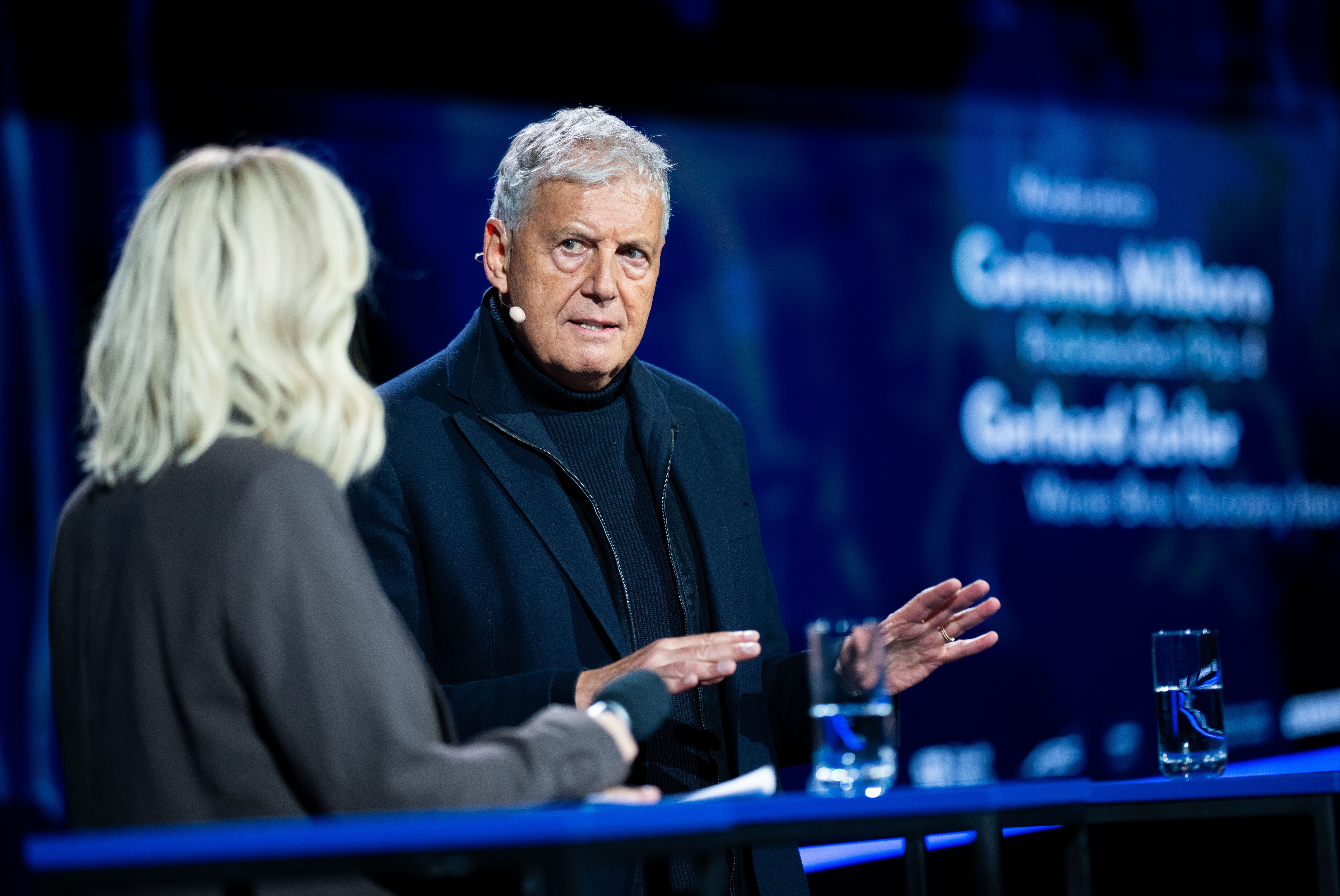 Zwei Personen sprechen bei einer Podiumsdiskussion, beide mit Mikrofonen, vor blauem Hintergrund.