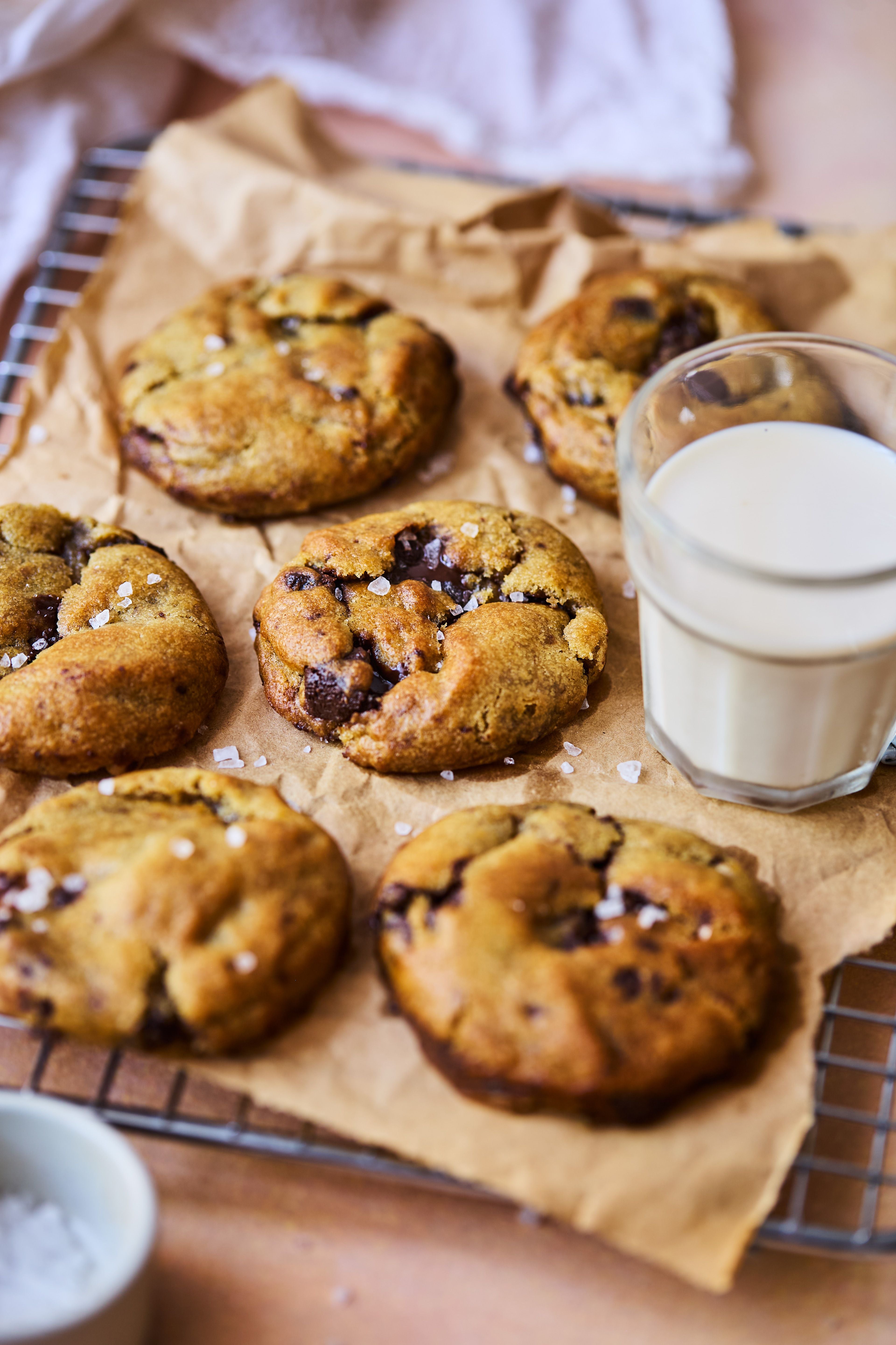 Sechs Chocolate Chip Cookies mit grobem Salz auf Backpapier, daneben ein Glas Milch.