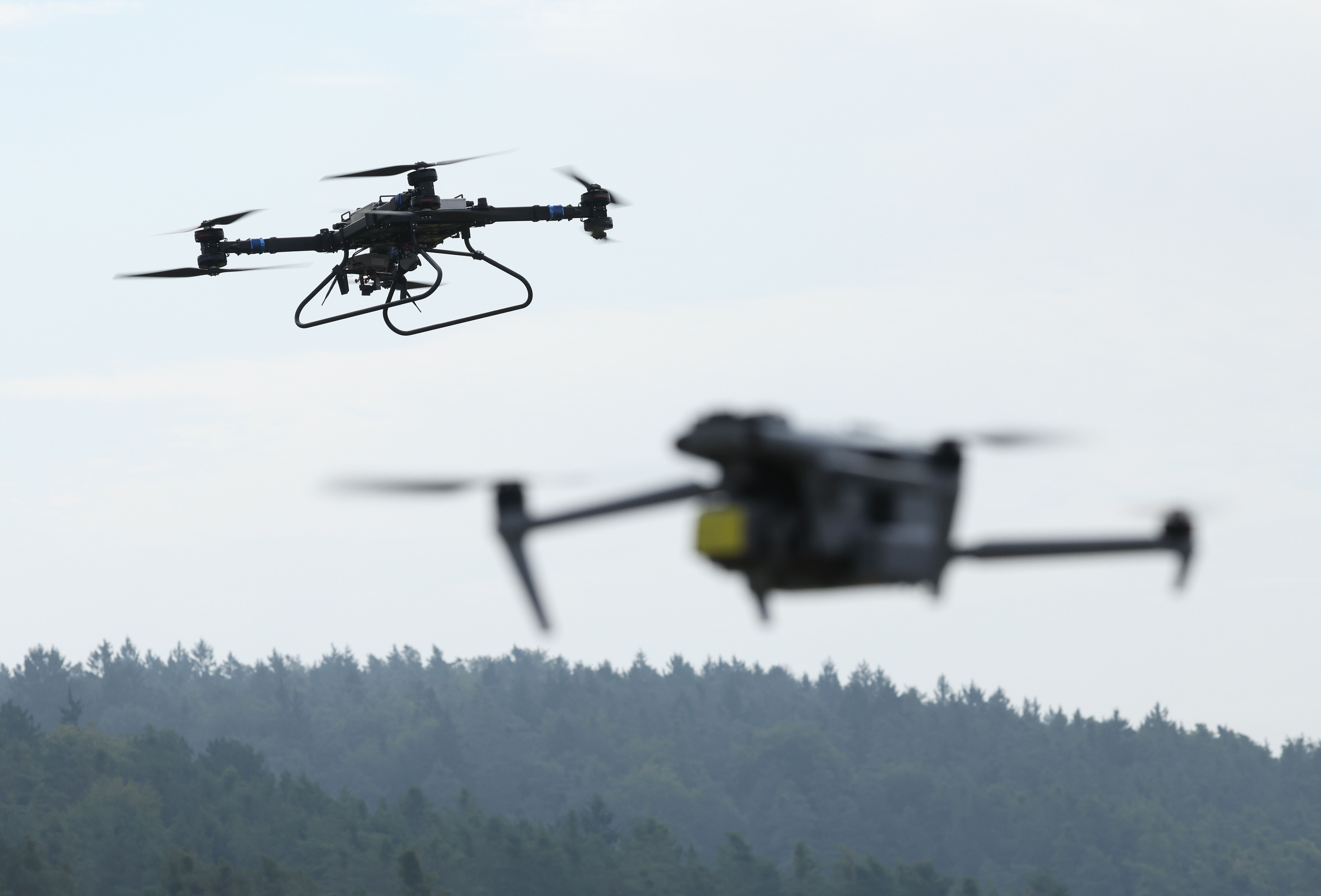 Zwei Drohnen fliegen in der Luft über einem bewaldeten Hügel bei Tageslicht.