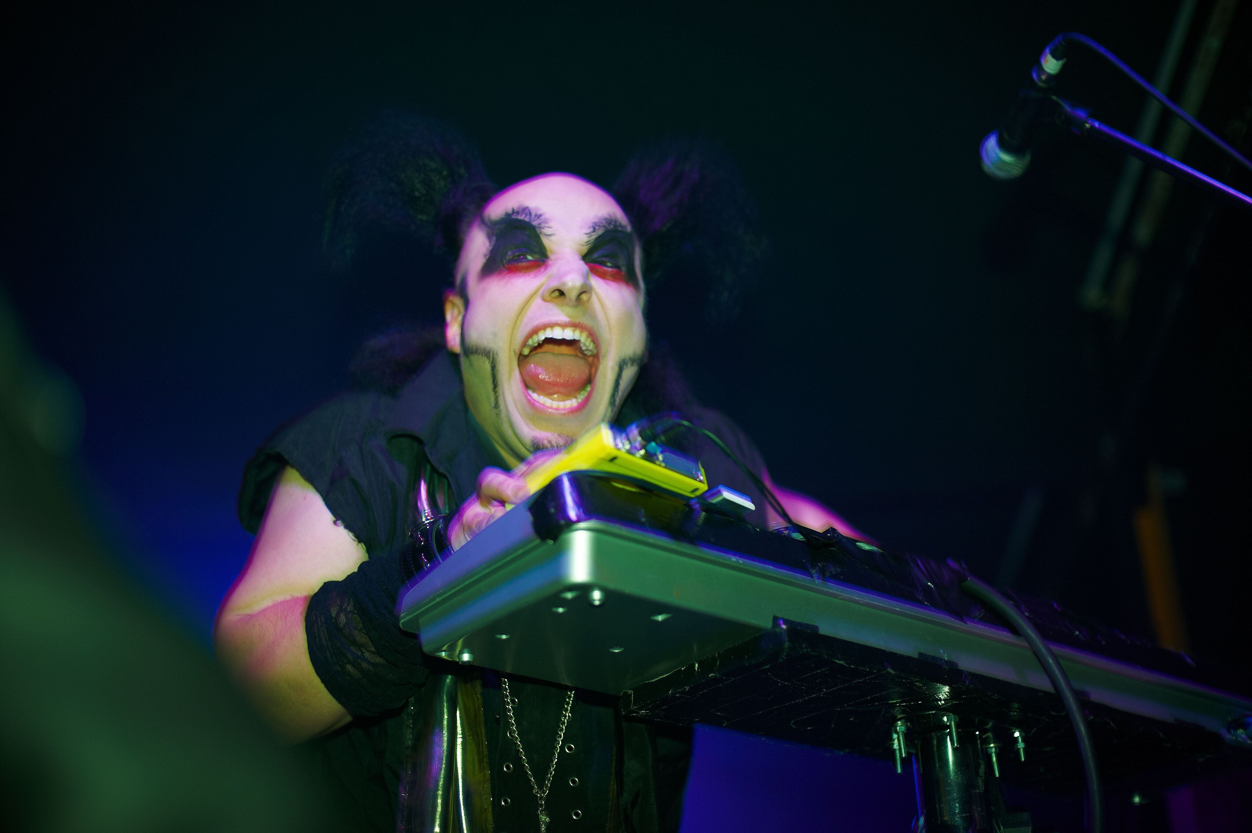 Ein Musiker mit auffälligem Make-up spielt Keyboard auf einer Bühne und schreit energetisch.