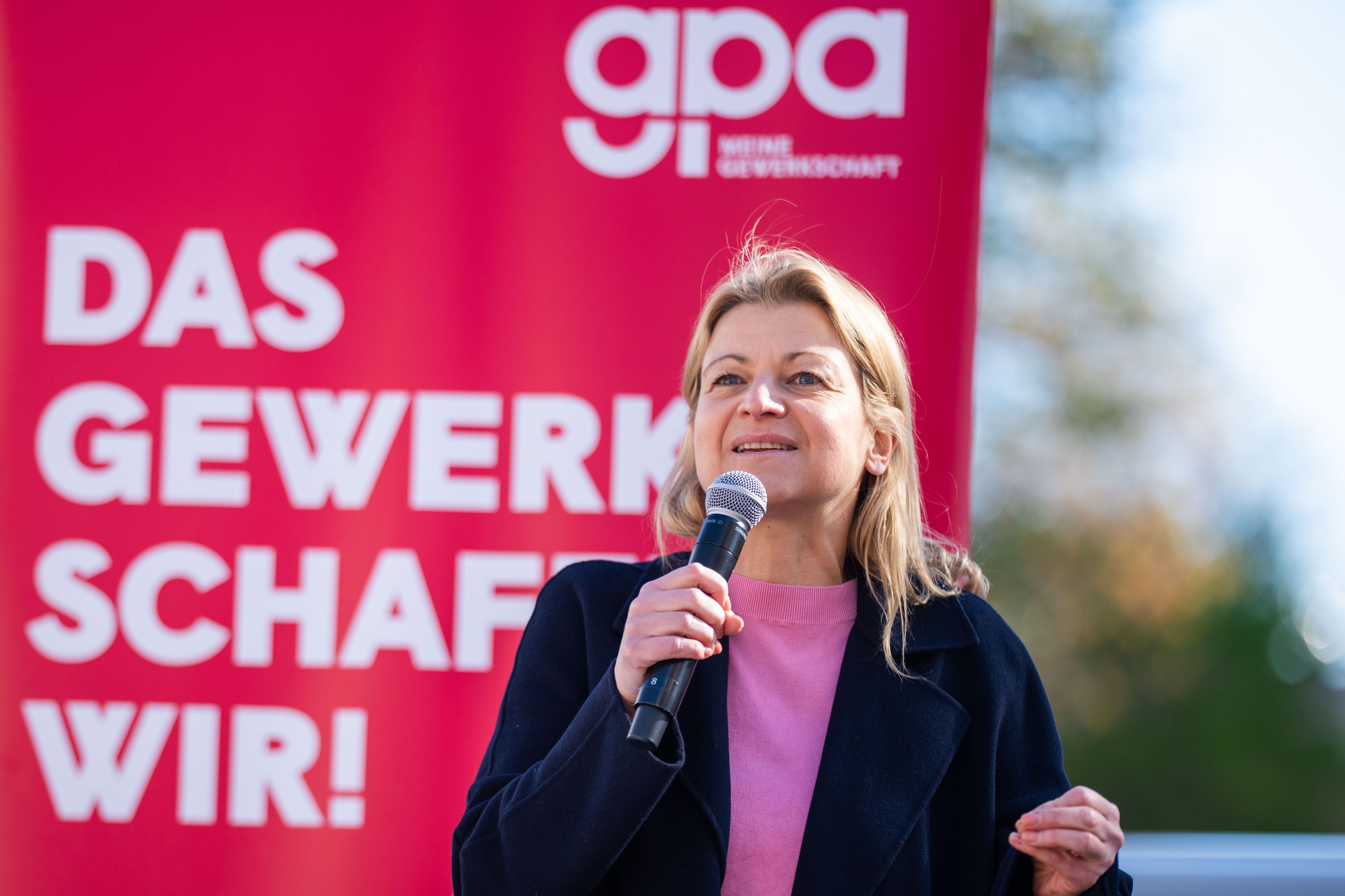 Eine Frau spricht bei einer Gewerkschaftsdemonstration mit Mikrofon vor rotem Banner.