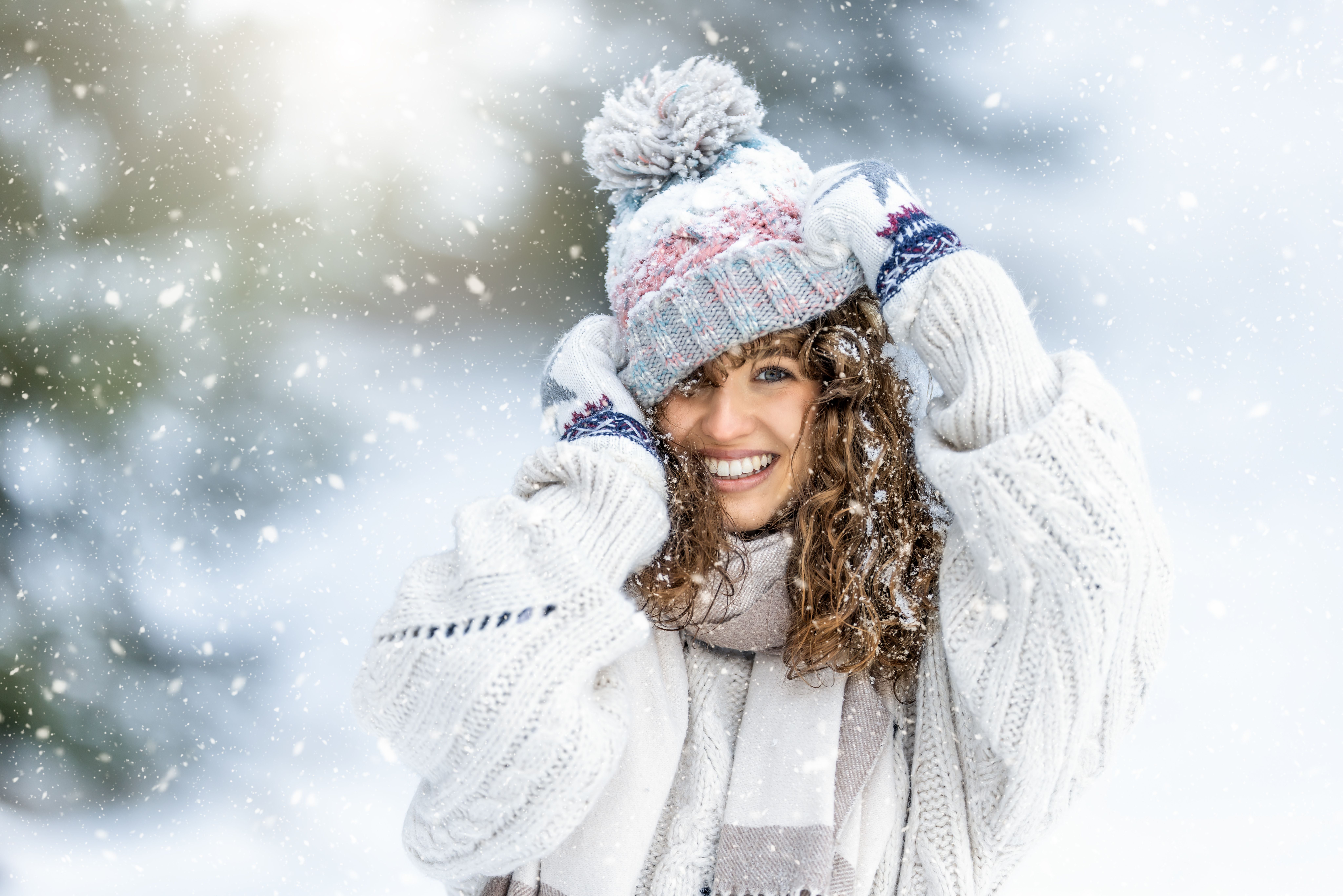 Junge Frau mit lockigem Haar lächelt im Schnee, trägt Strickmütze, Schal und Handschuhe.