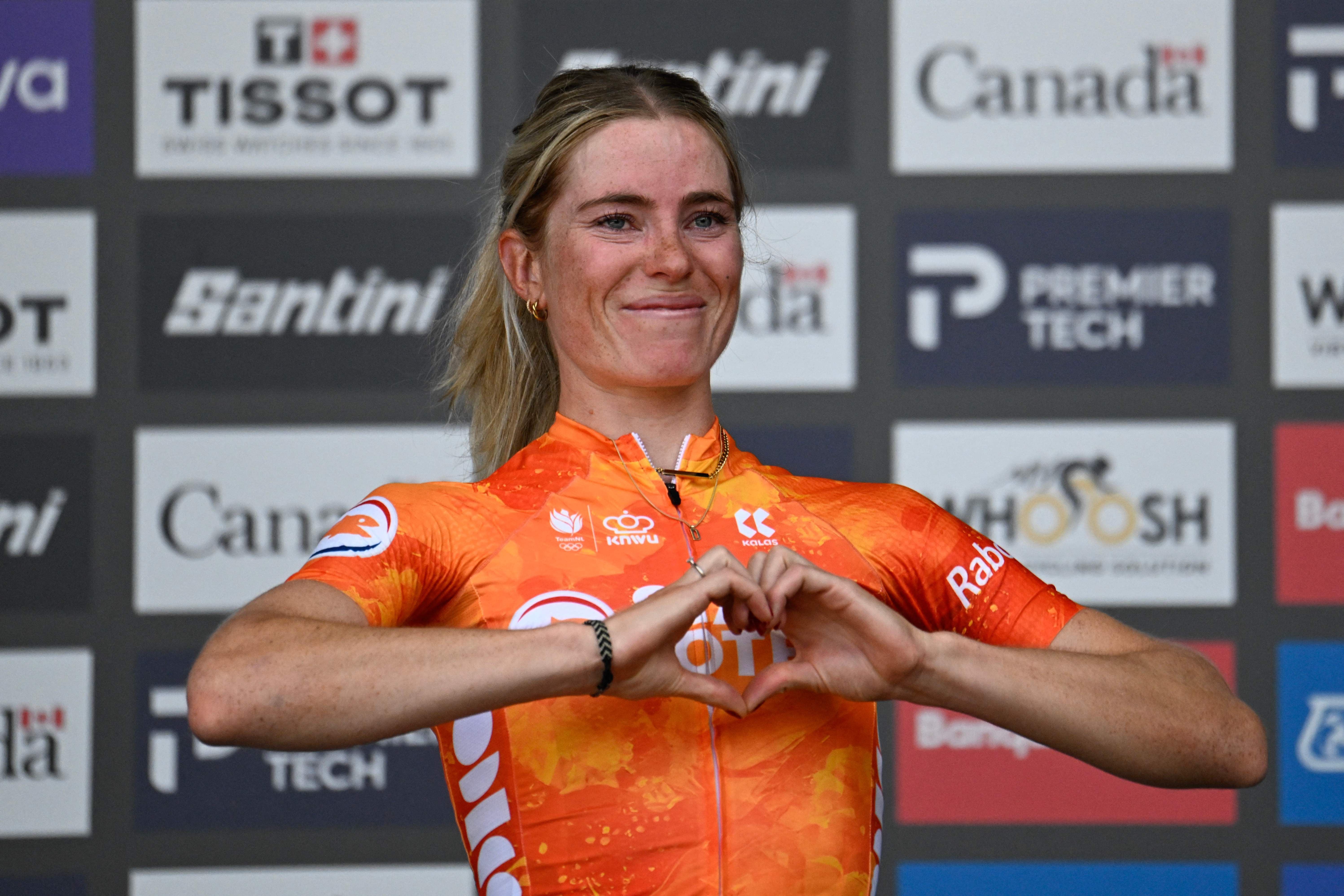 Eine Radrennfahrerin im orangefarbenen Trikot formt auf dem Podium ein Herz mit den Händen.